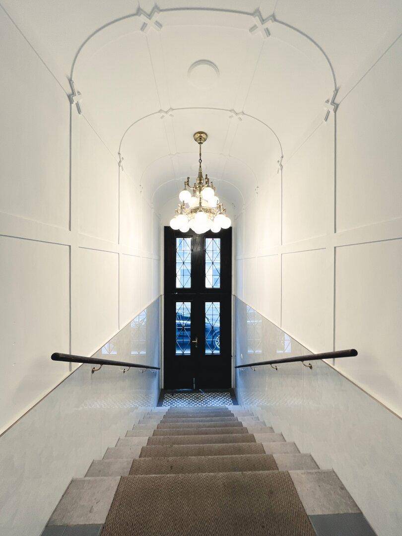 Historisches Treppenhaus mit eleganten Stufen, hohen Decken und einer verzierten Eingangstür.