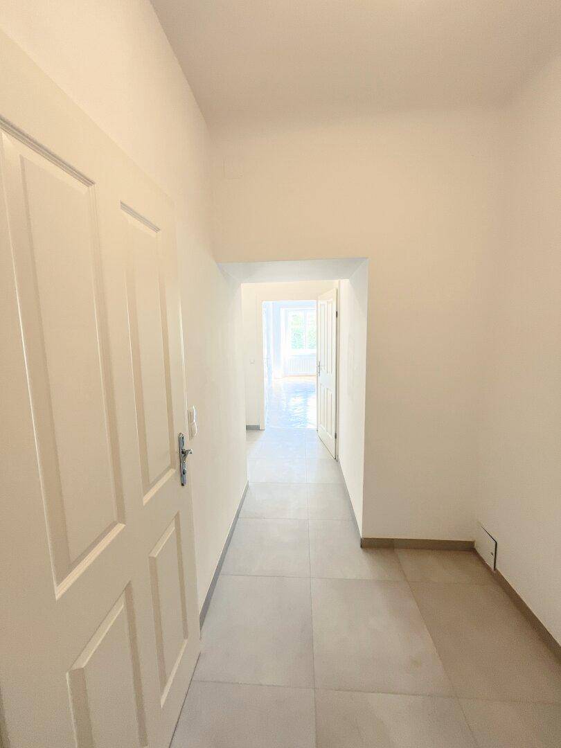 Geräumiger Flur mit Fliesenboden und Zugang zu einem hellen Bereich im Hintergrund.