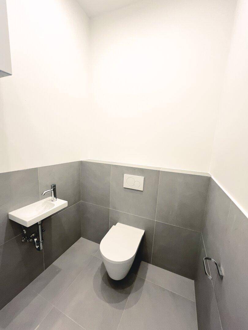 Modernes Gäste-WC mit grauen Fliesen, wandhängendem WC und kleinem Waschbecken.