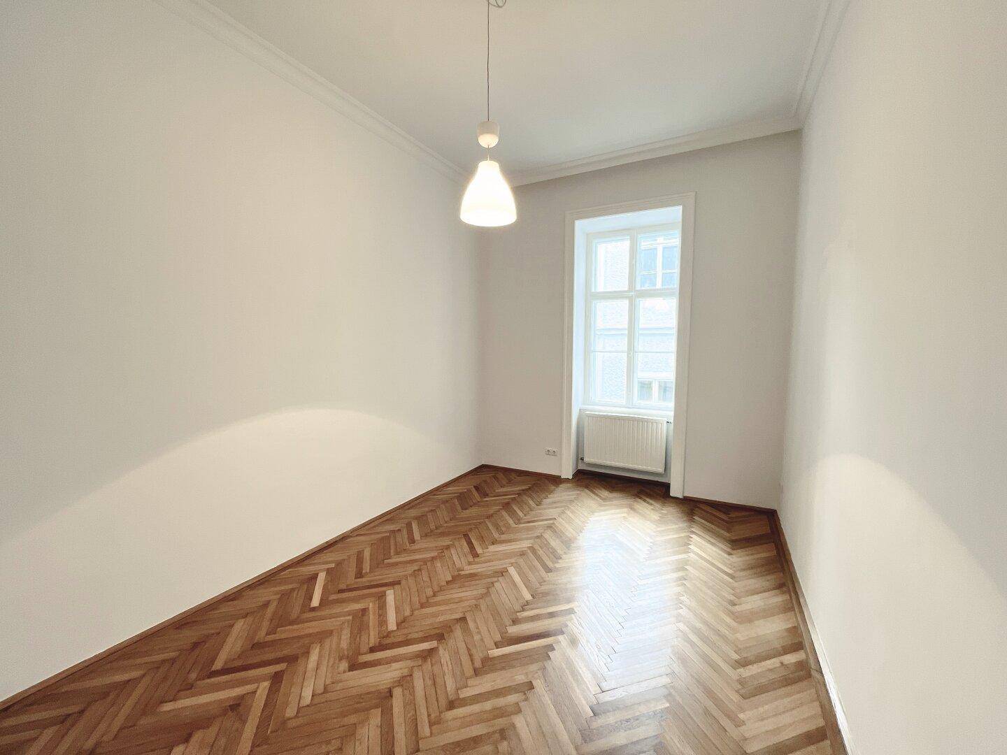 Helles Zimmer mit Fischgrätparkettboden, hohen Decken und einem großen Fenster.
