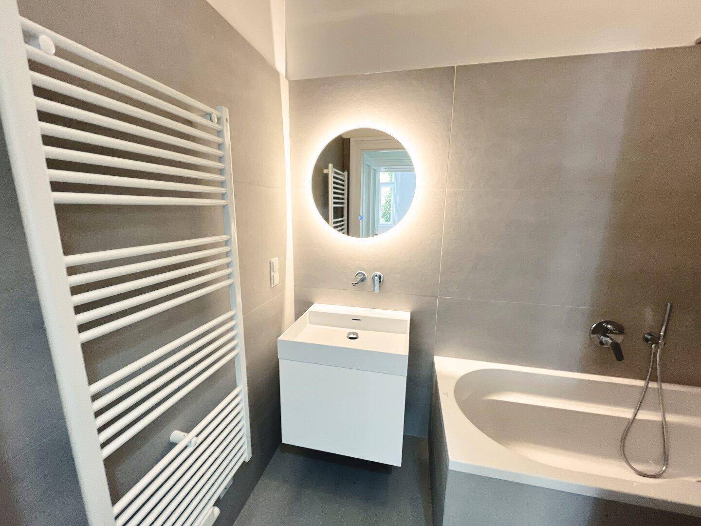Zeitgemäßes Badezimmer mit Badewanne, Waschtisch und beleuchtetem Wandspiegel.