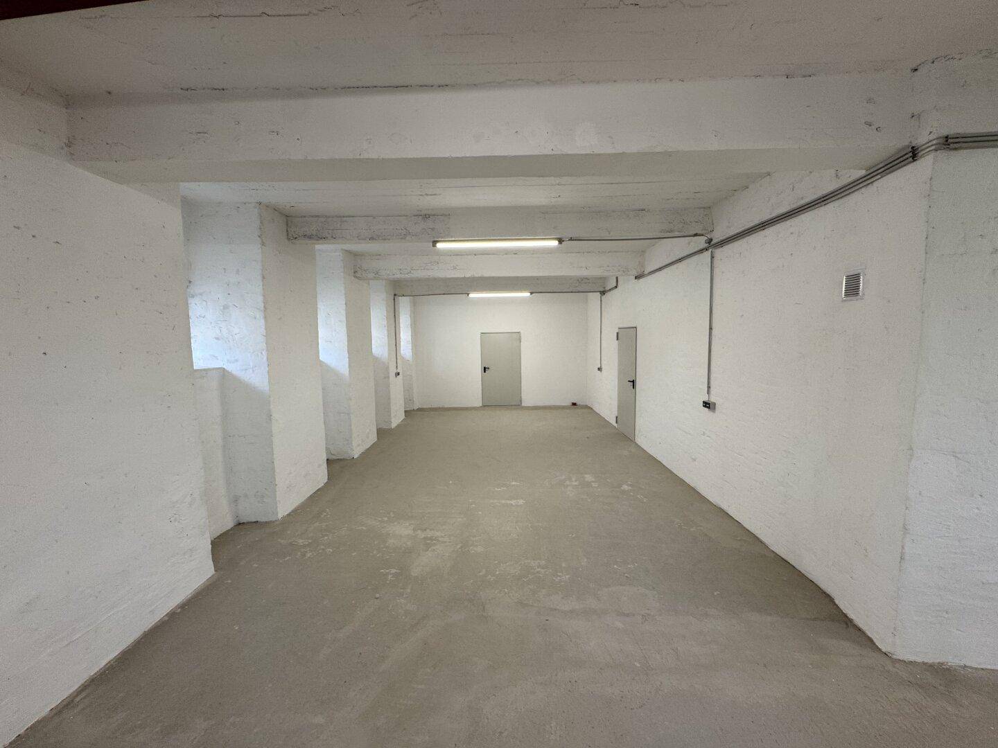 Langer, leerer Lagerraum mit weißen Wänden, Betonboden und Deckenbeleuchtung.
