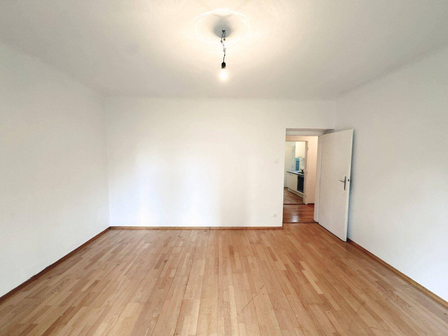 Heller Raum mit Parkettboden und Zugang zu einer Küche, ideal als Wohnzimmer.