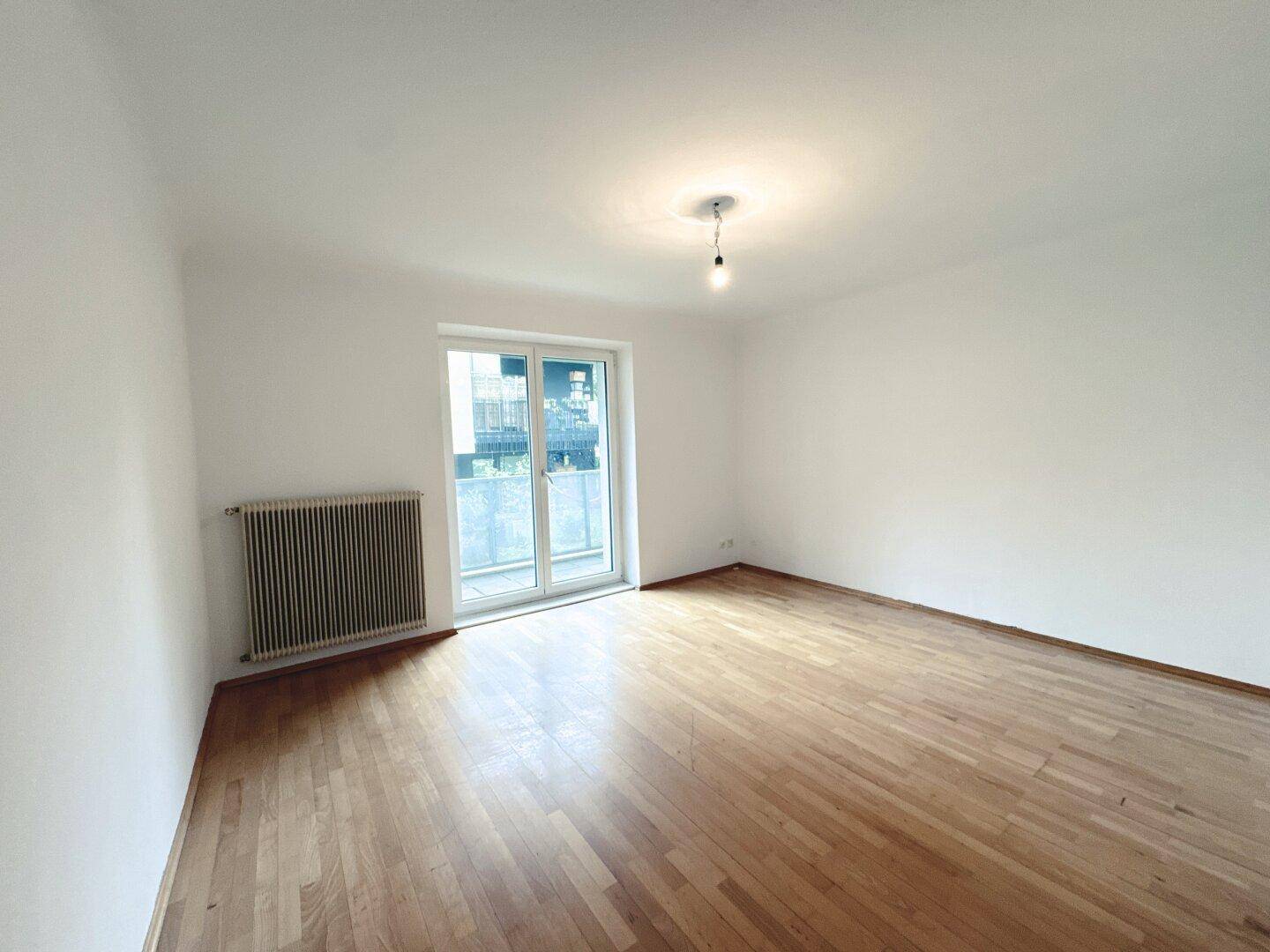 Helles, leeres Wohnzimmer mit Parkettboden und Zugang zu einem Balkon.