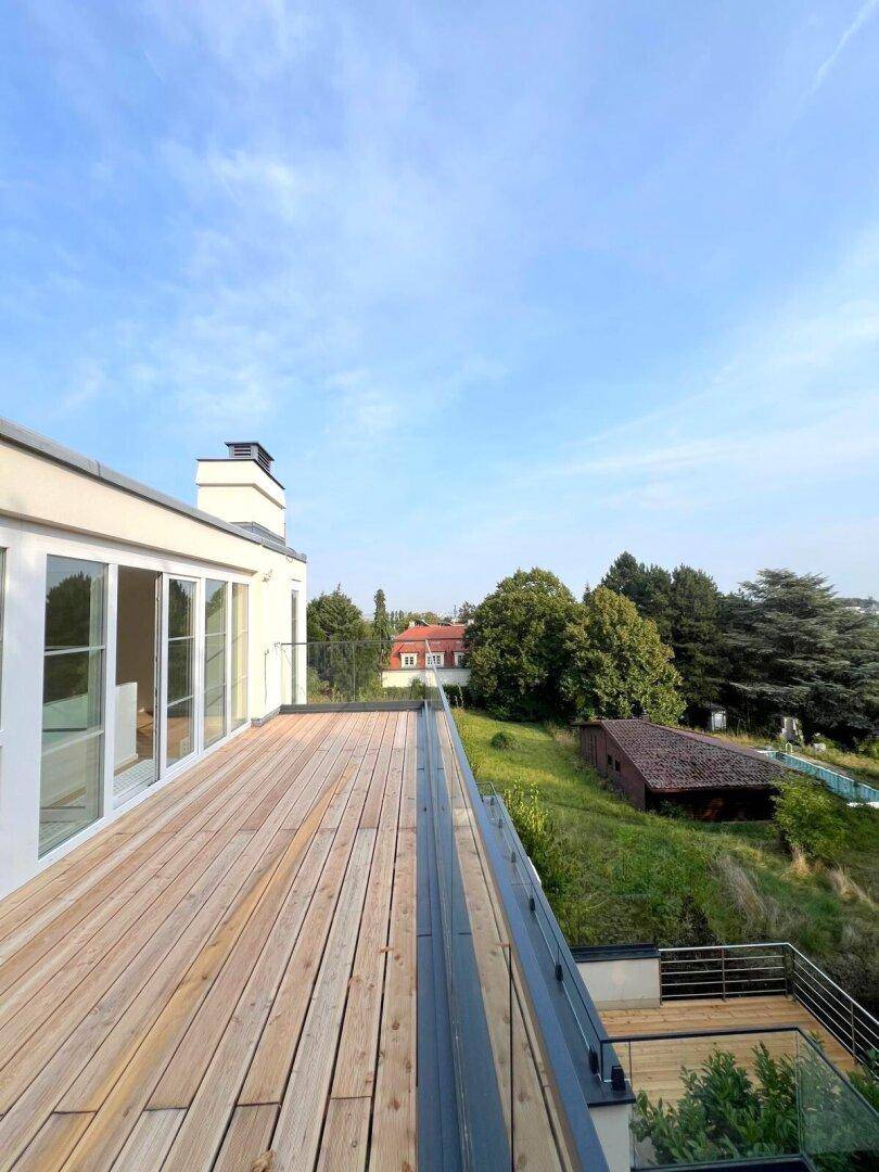 Weitläufige Holzterrasse mit Glasgeländer und Blick auf die grüne Umgebung.