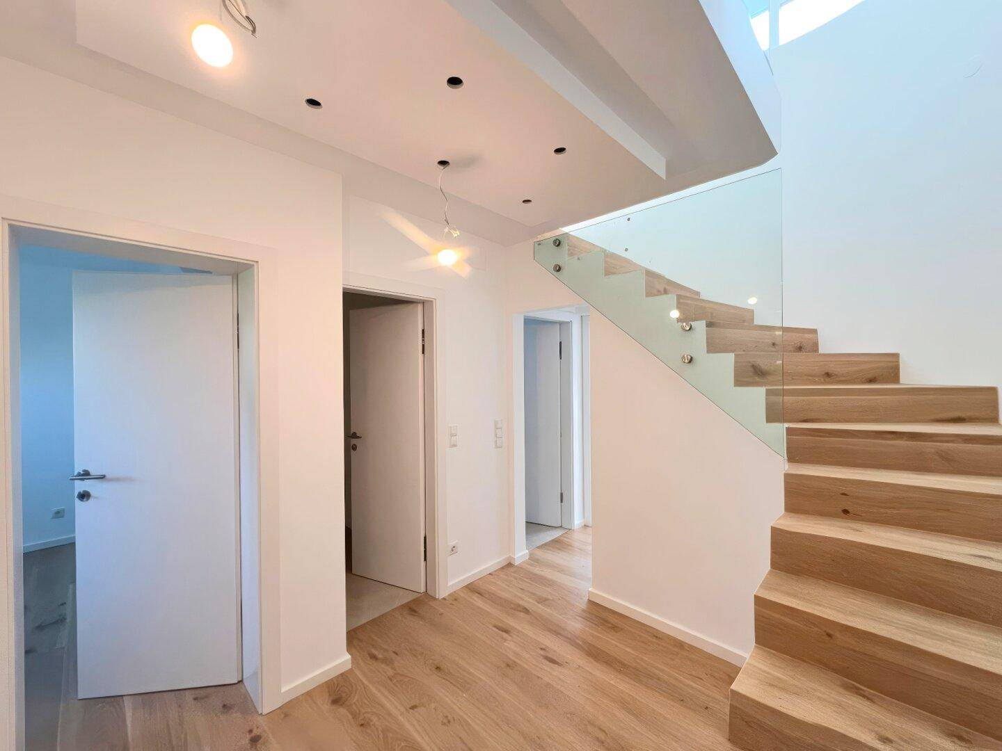 Heller Flur mit moderner Holztreppe und Glasgeländer, der zu verschiedenen Räumen führt.