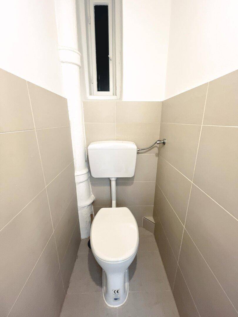 Separates WC mit Toilette und kleinem Fenster, halbhoch gefliest in neutralen Farben.