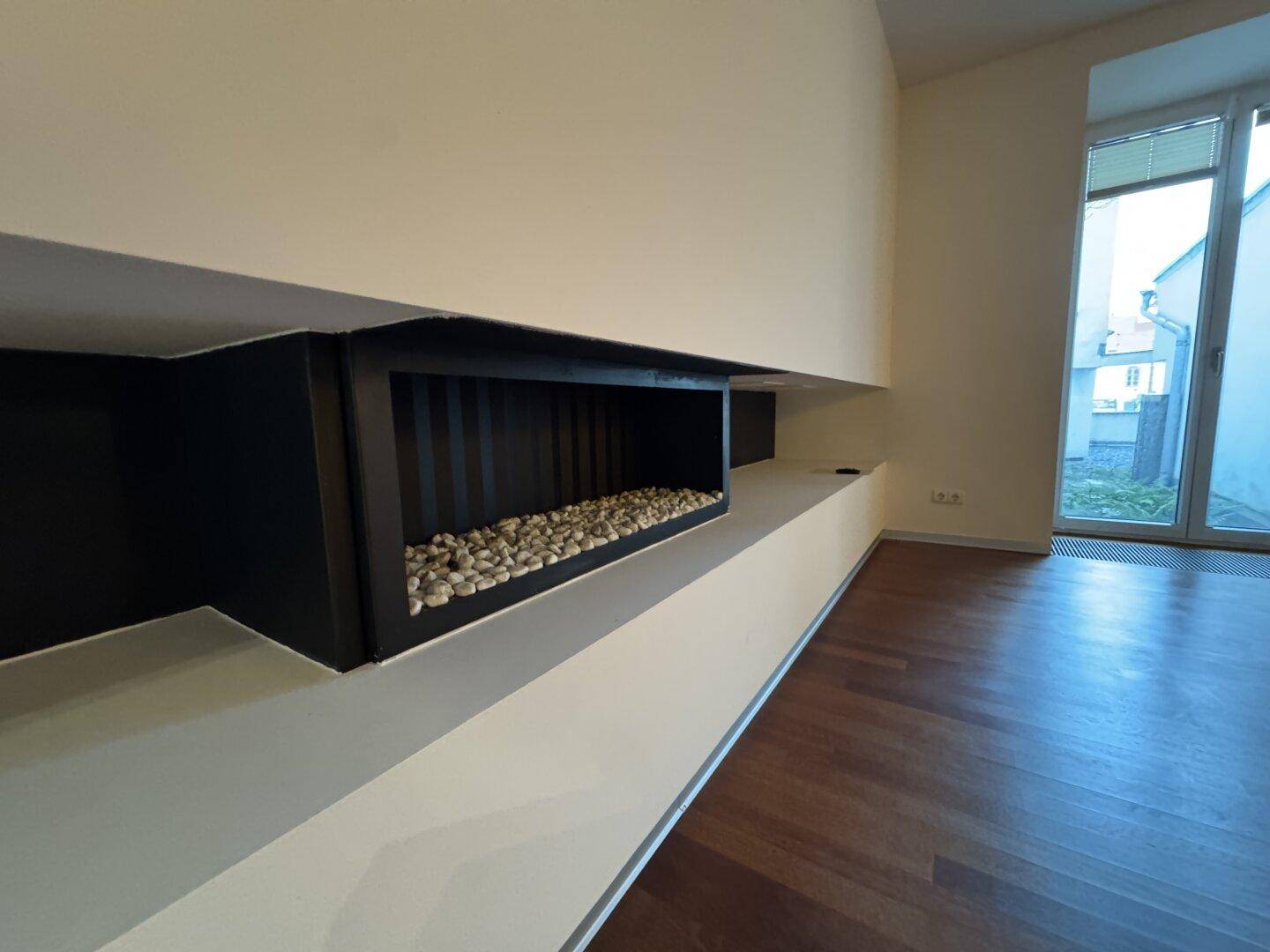 Moderner Kamin mit dekorativen Steinen, elegant in die Wand integriert, ergänzt durch Holzboden und Fenster.