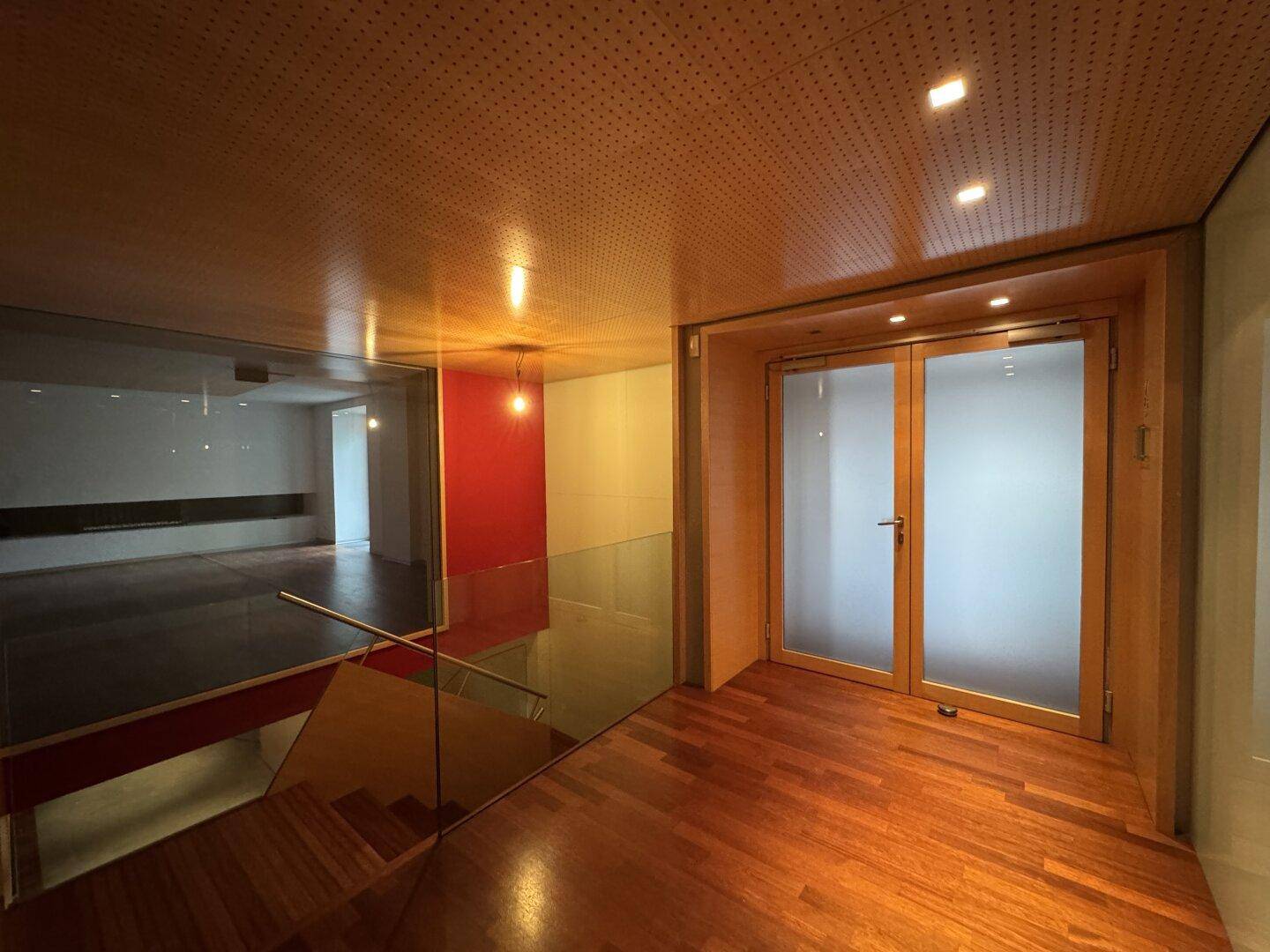 Heller Flur mit Holzboden, Glasgeländer und satinierten Glastüren, unter einer perforierten Holzdecke.