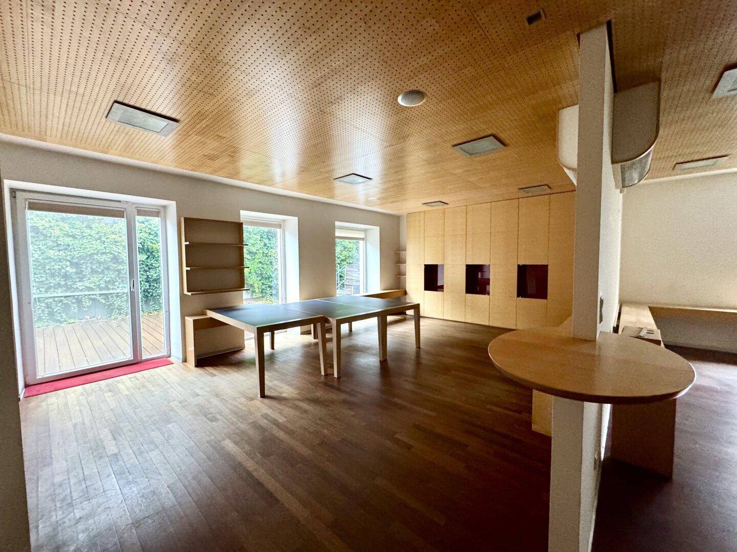 Großer Mehrzweckraum mit Holzboden, perforierter Holzdecke, Einbauschränken und Zugang zu einem Außenbereich.