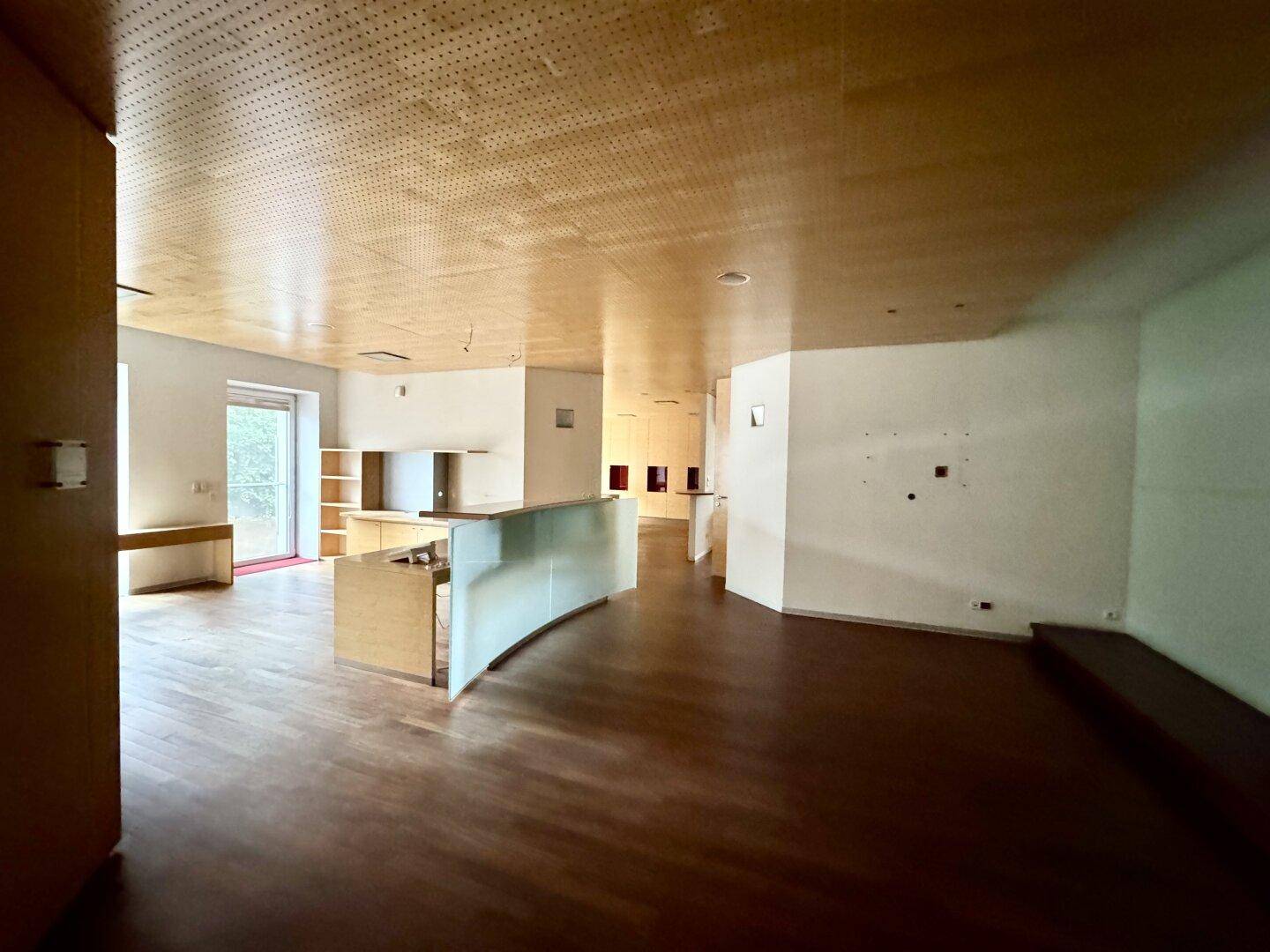 Geräumiger Mehrzweckraum mit Holzboden, perforierter Holzdecke und einer modernen, geschwungenen Theke.
