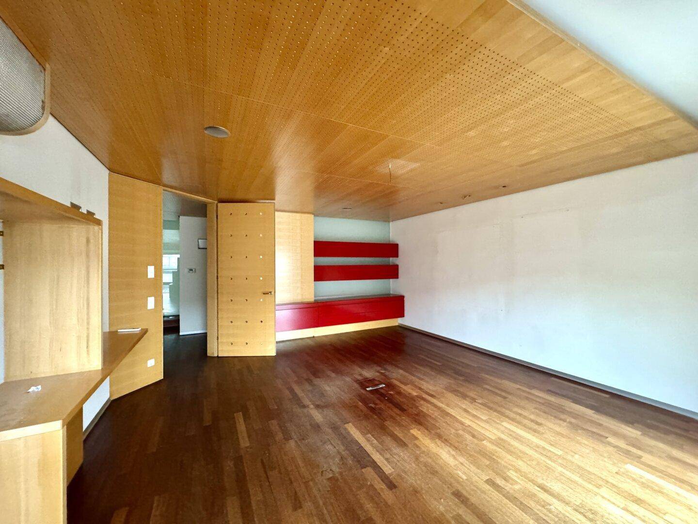 Heller Raum mit Holzboden, perforierter Holzdecke und einer roten Akzentwand mit modernen Regalen.