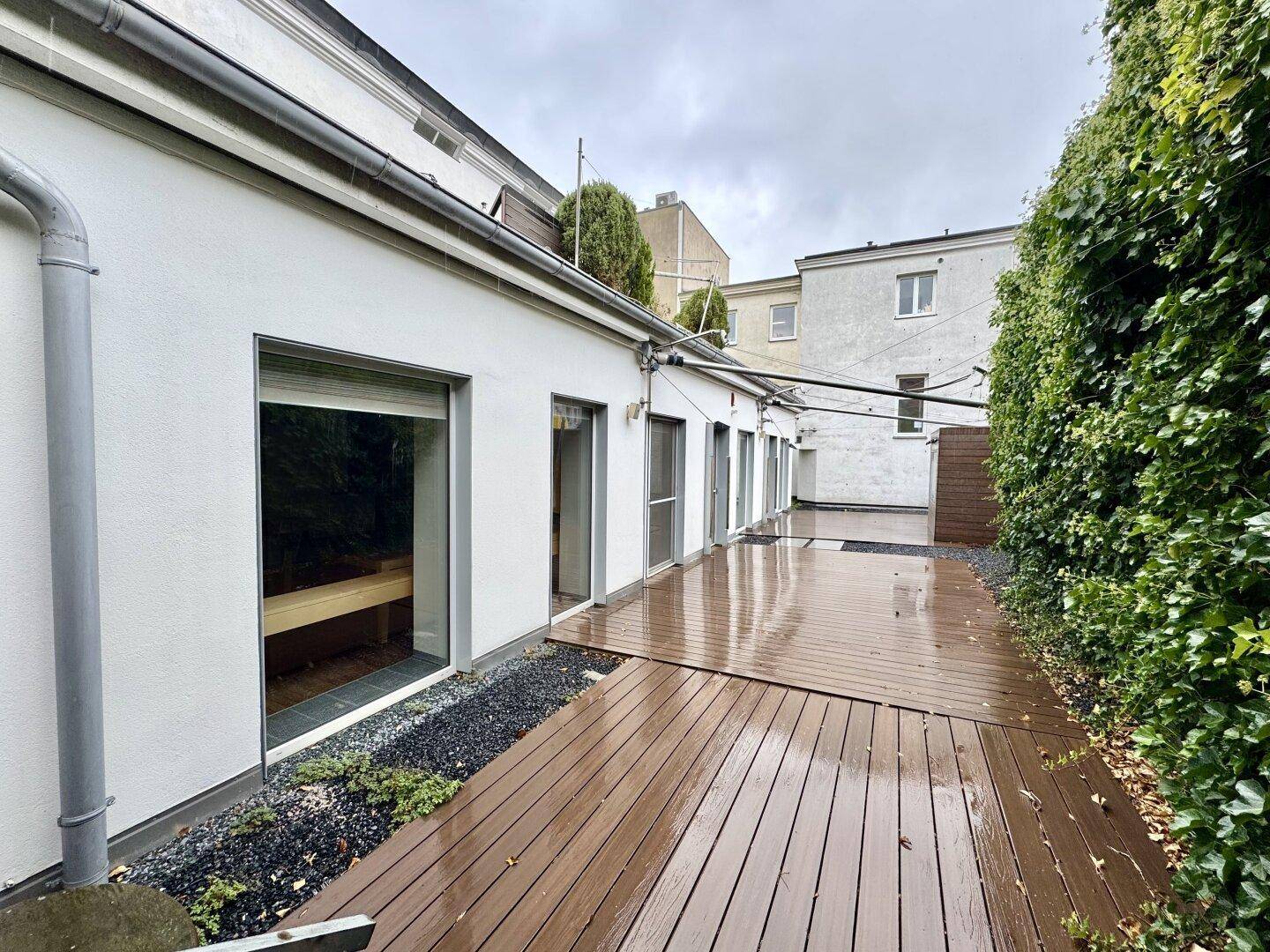 Blick auf die Terrasse mit Holzdielen, umgeben von einer grünen Hecke und der Gebäudefassade mit mehreren Zugängen.