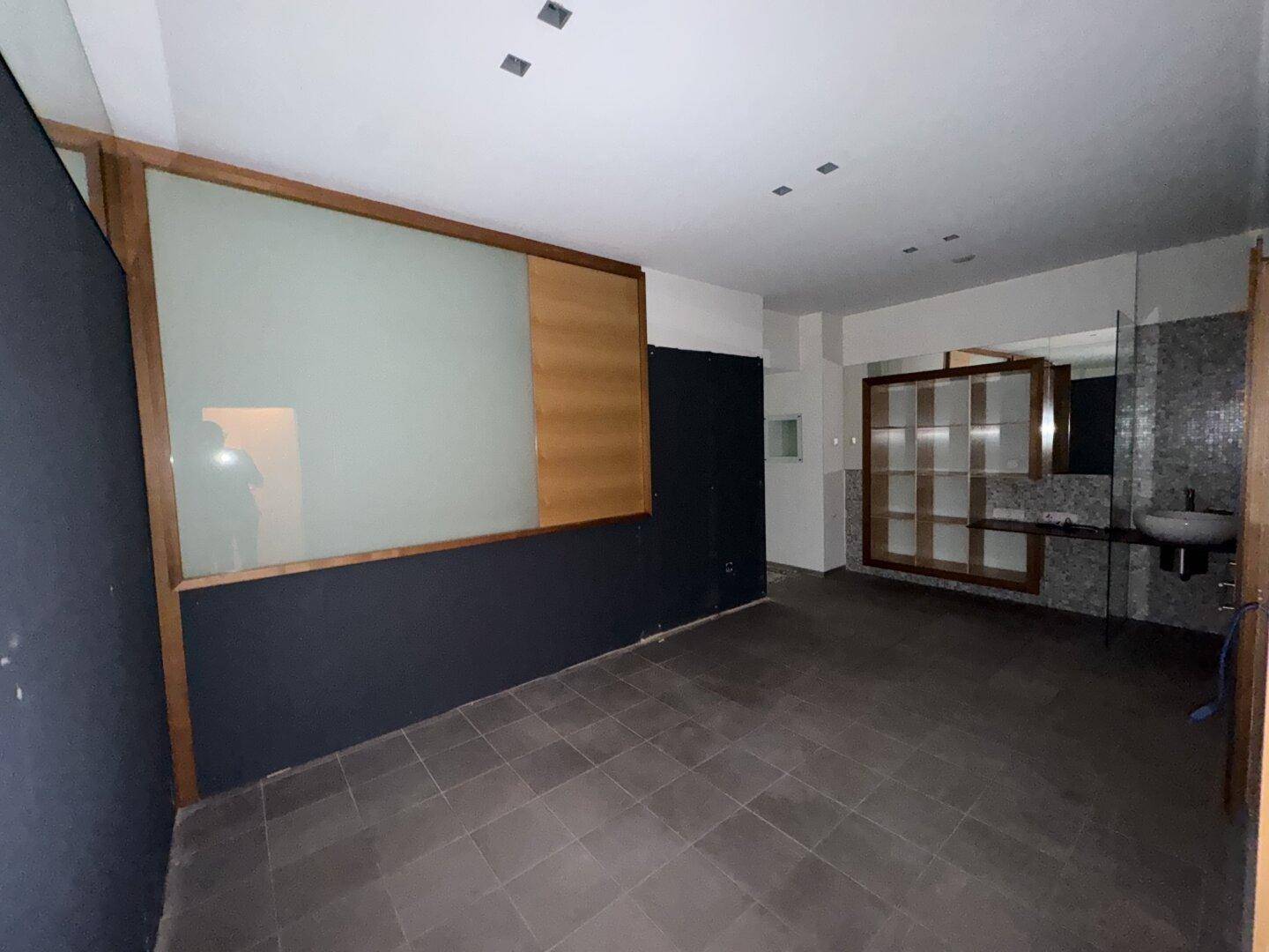 Raum mit dunklem Fliesenboden, einem Waschbecken und einer Glasabtrennung, möglicherweise ein Badezimmer oder Umkleidebereich.