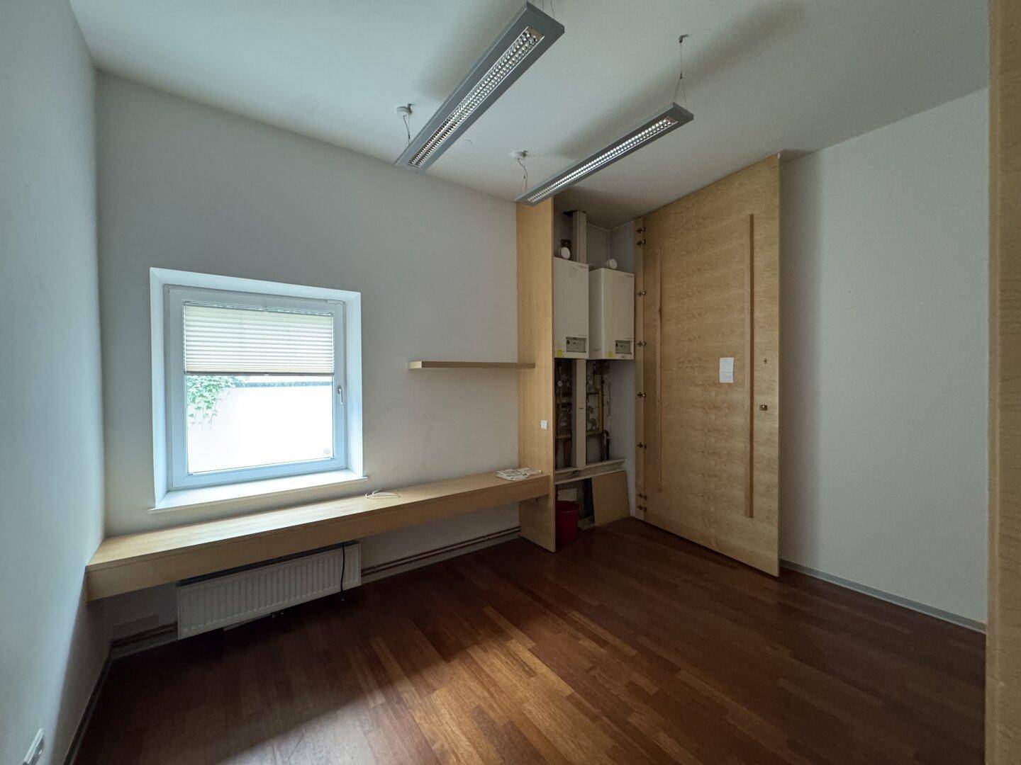 Kleiner Raum mit Holzboden, Fenster und einer integrierten Holzbank, dahinter ist die Heizungsanlage sichtbar.