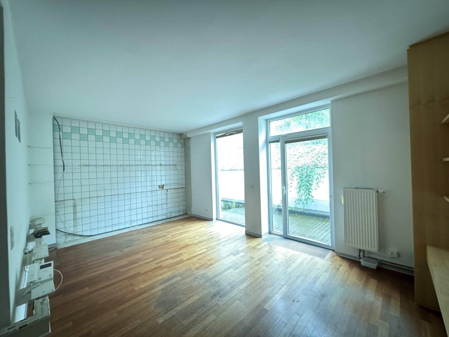 Unmöblierte Küche mit gefliesten Wänden, Holzboden und Zugang zur Terrasse durch große Glastüren.