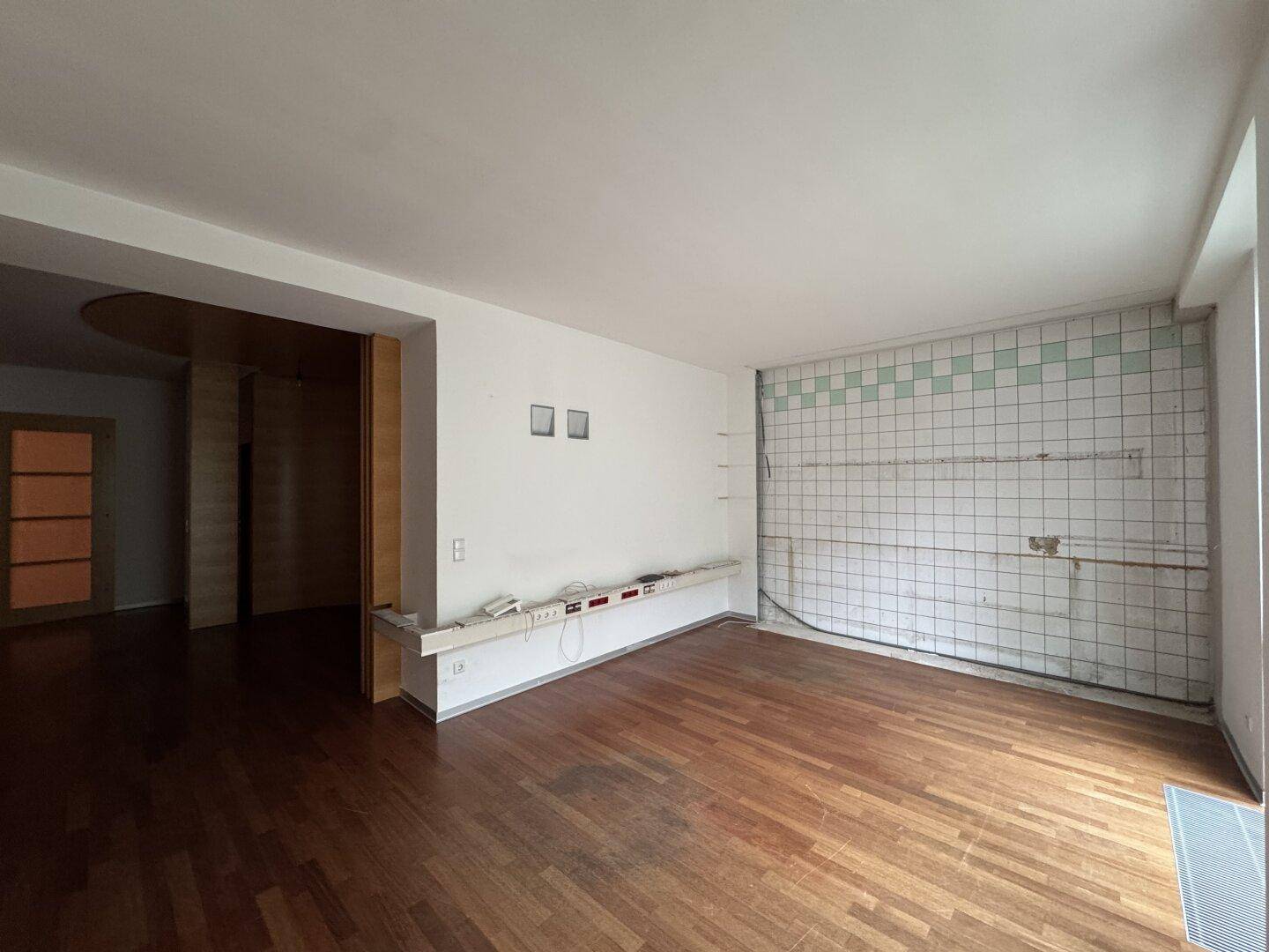 Raum mit Holzboden, weißen Wänden und einem gefliesten Bereich an einer Wand, möglicherweise von einer ehemaligen Küche.