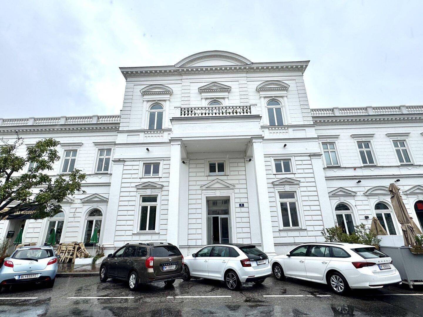 Prächtiges weißes Gebäude mit klassischer Architektur und geparkten Autos davor an einem bewölkten Tag.