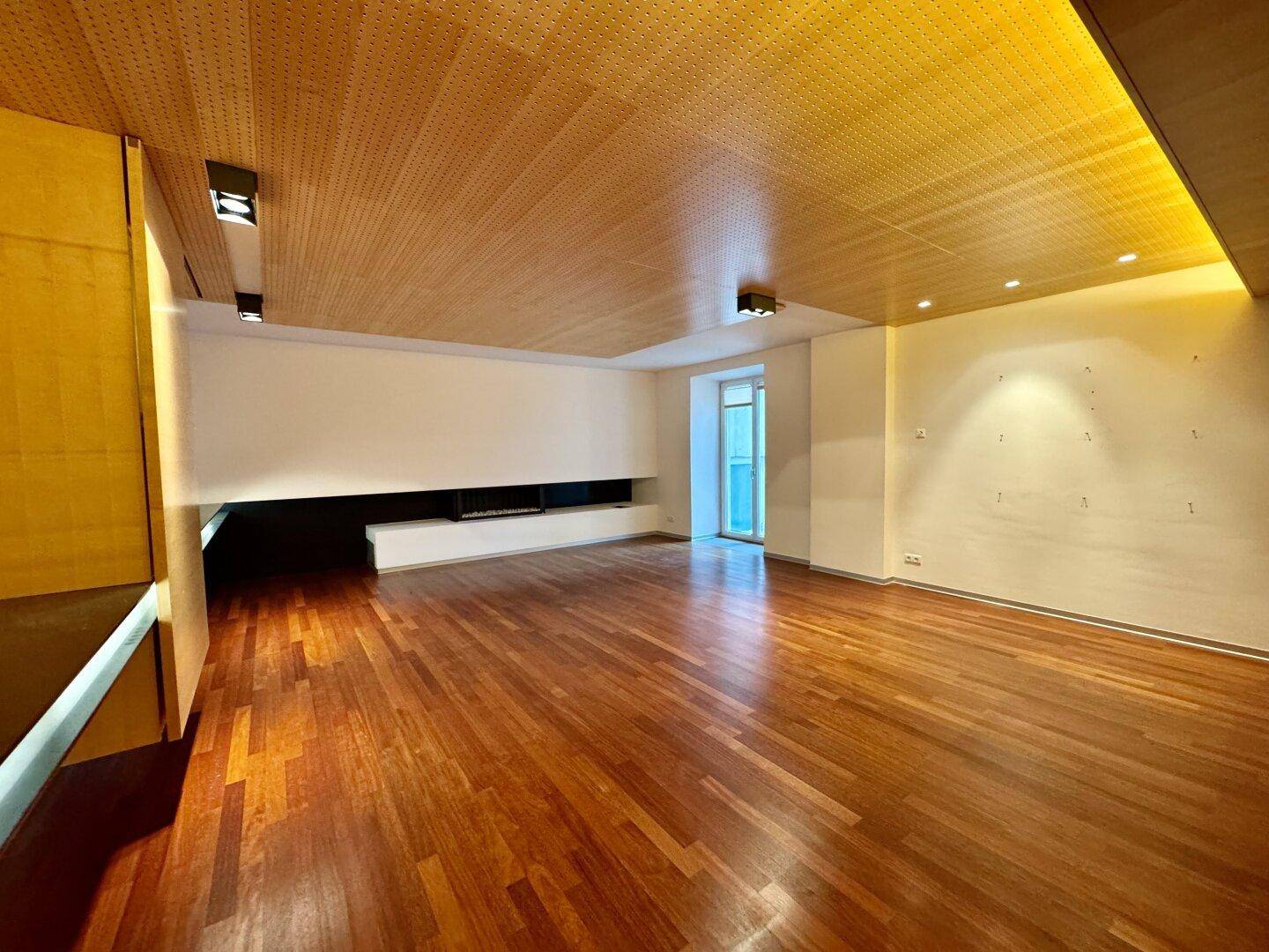 Geräumiger, leerer Wohnraum mit Holzboden, perforierter Holzdecke und einem langen, integrierten Wandelement.