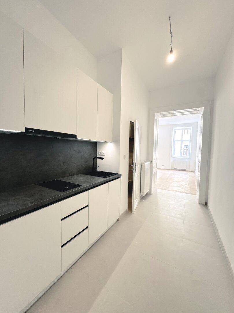 Schmale, moderne Küche mit weißen Schränken, dunkler Arbeitsplatte und Blick in einen weiteren Raum.