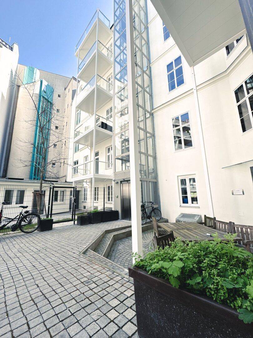 Innenhof mit modernem Glasaufzug, Sitzgelegenheiten und begrünten Pflanzkübeln.