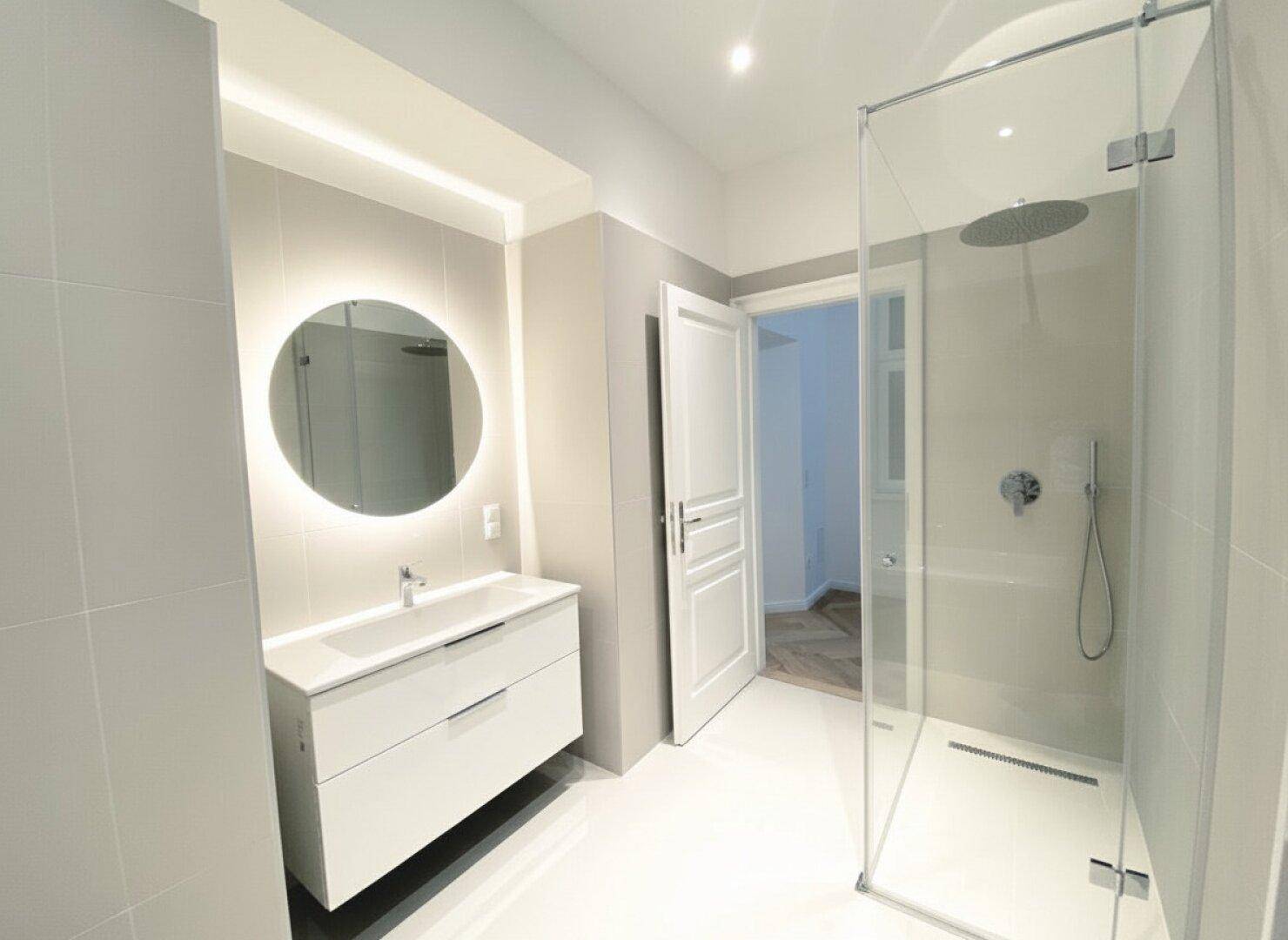 Modernes Badezimmer mit begehbarer Dusche, schwebendem Waschtisch und beleuchtetem Spiegel.