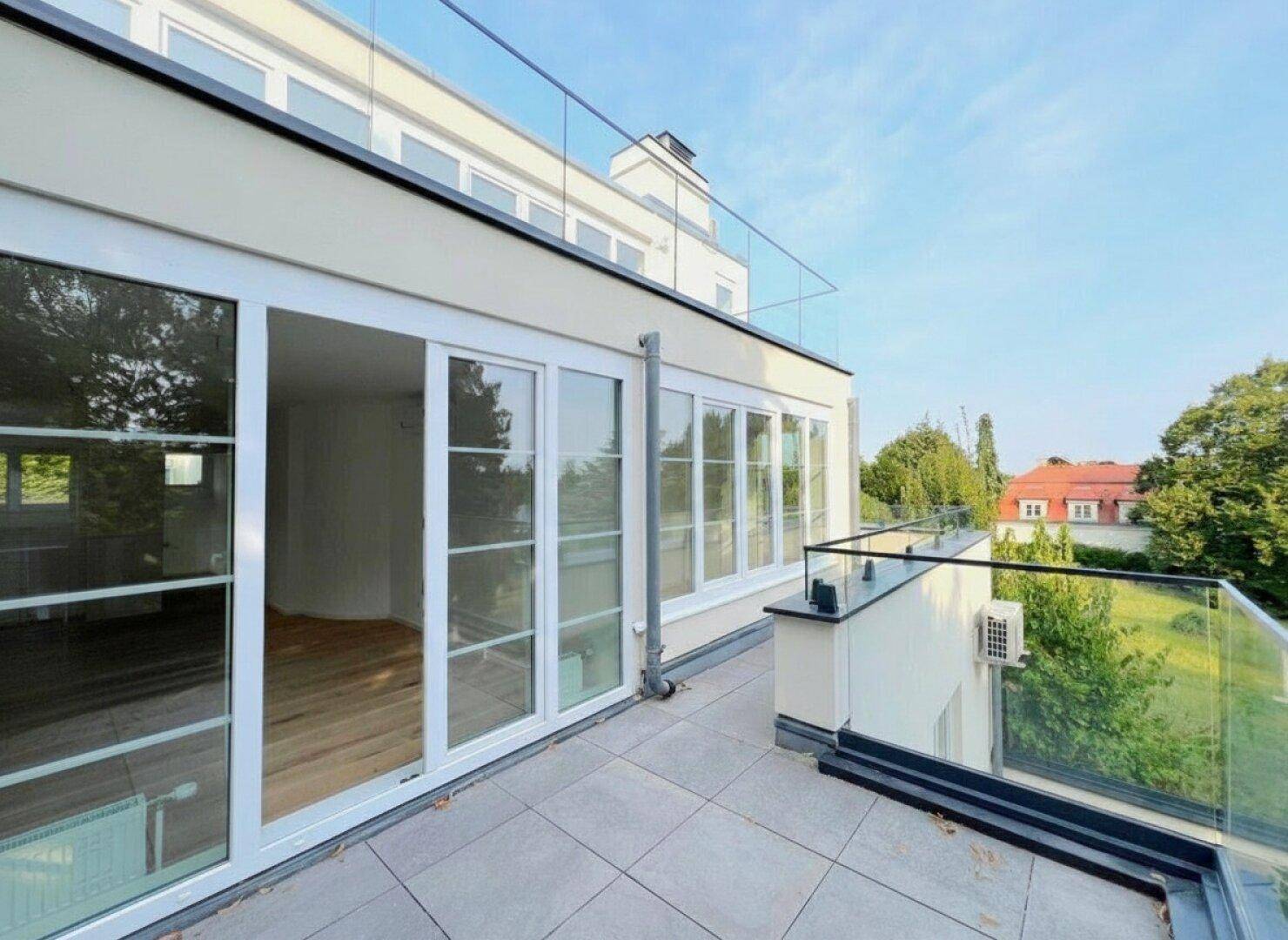 Geräumige Terrasse mit Fliesenboden, Glasgeländer und weitem Blick ins Grüne unter blauem Himmel.