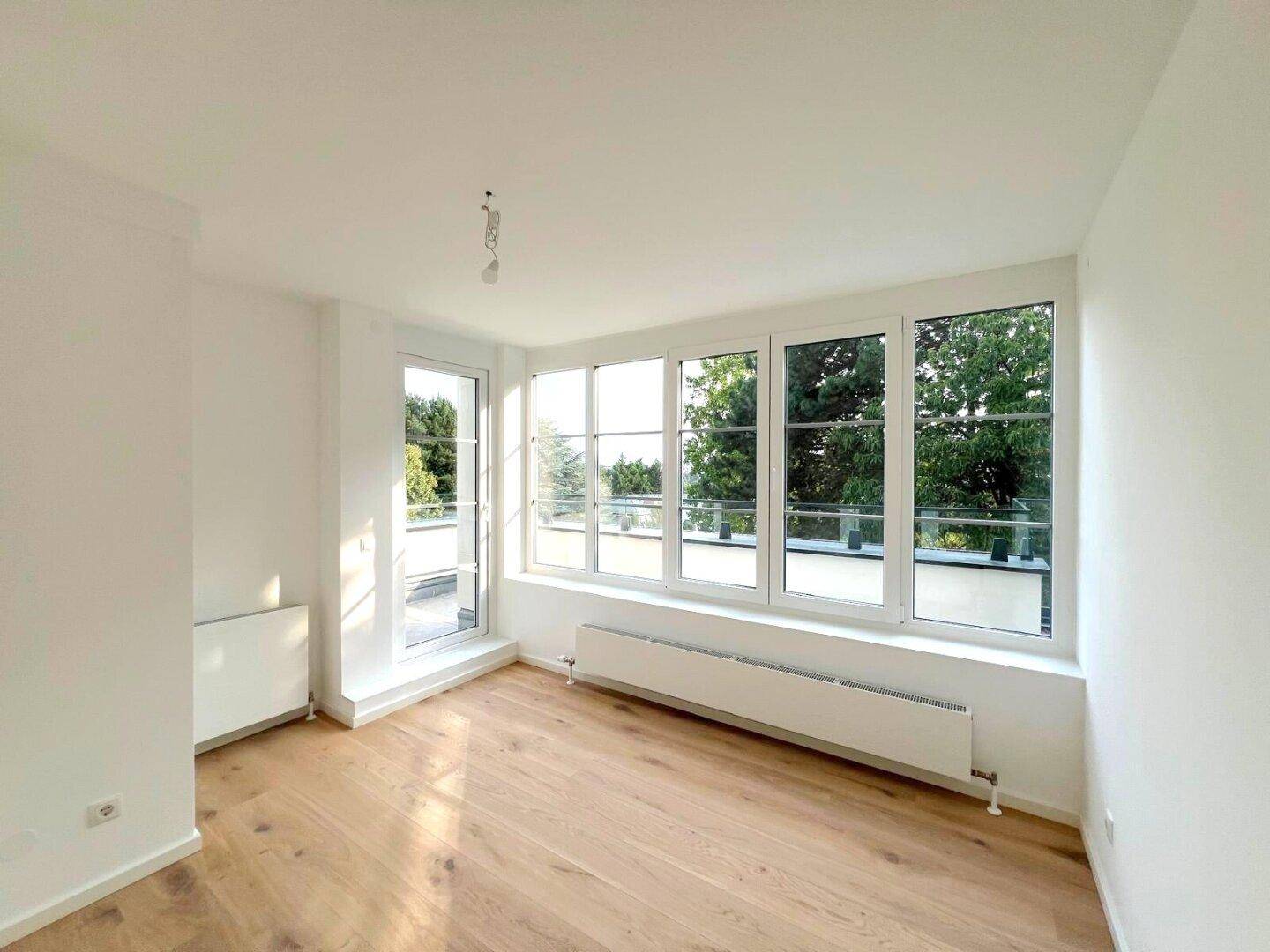 Heller Raum mit großen Fenstern, Holzboden und Blick ins Grüne.