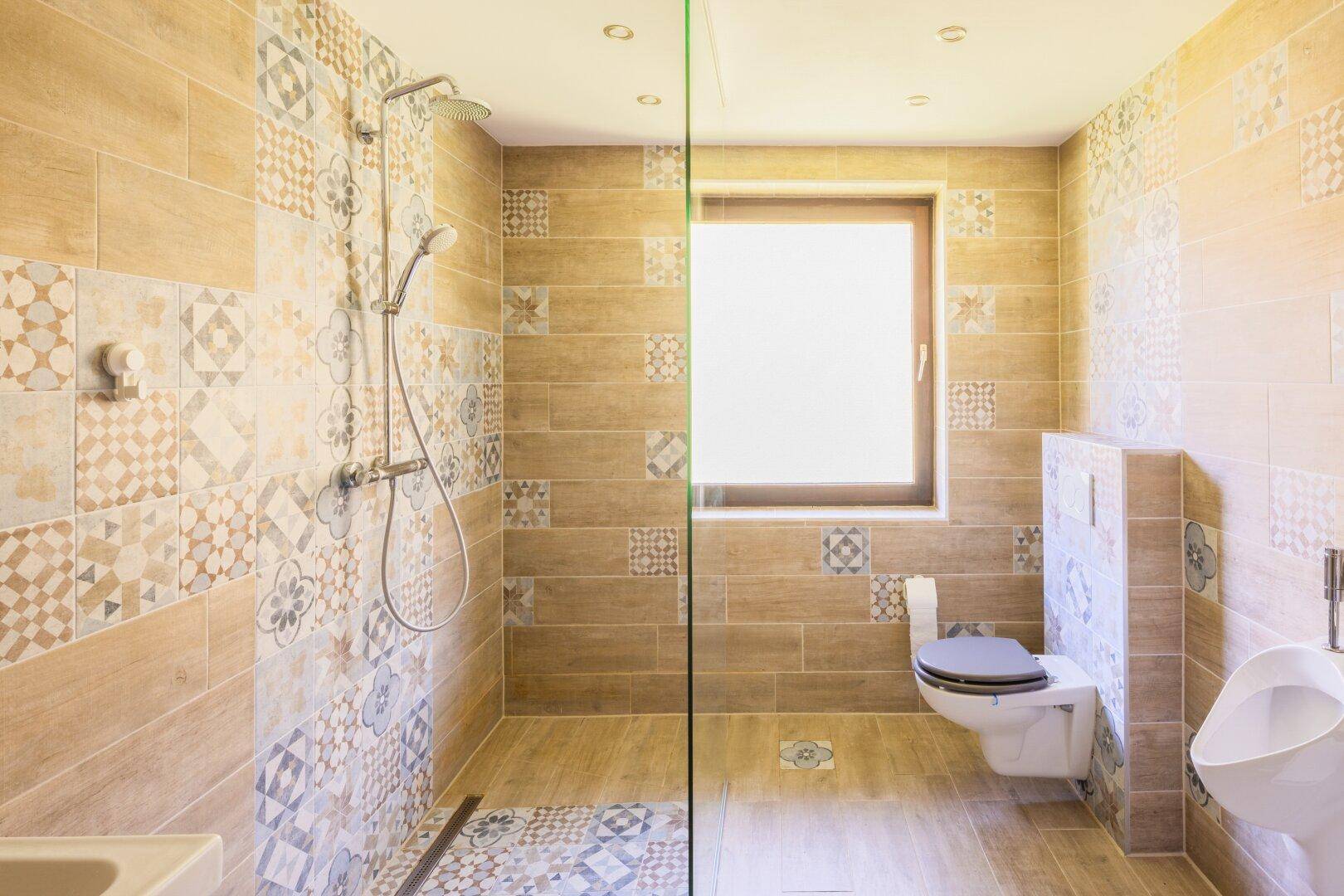 Geräumiges Badezimmer mit begehbarer Dusche, WC und Urinal, hell gefliest.