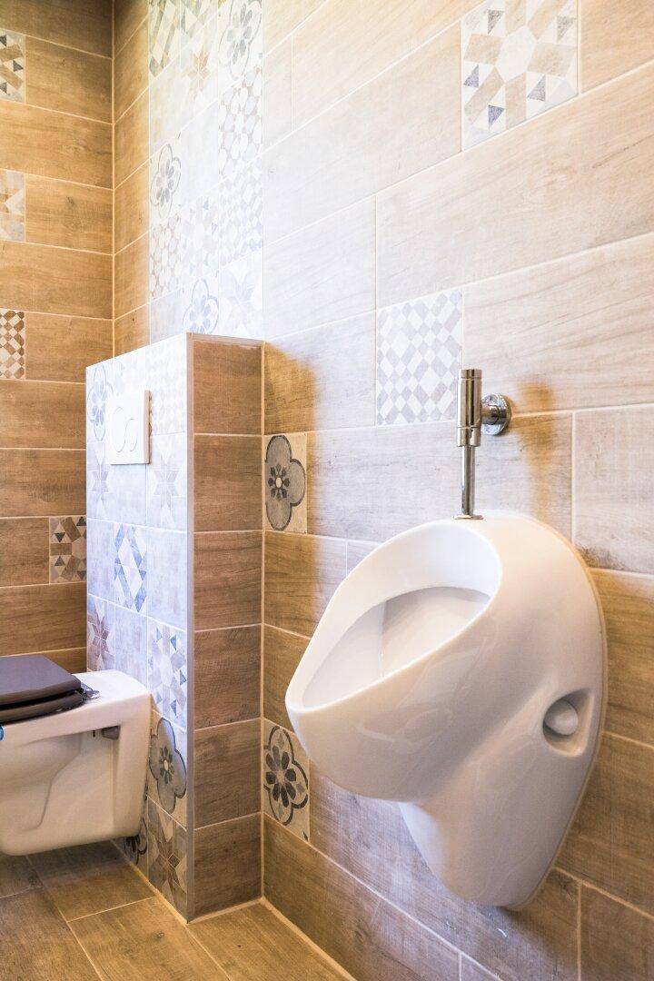 Badezimmerbereich mit Urinal, wandhängendem WC und gemusterten Fliesen an den Wänden.