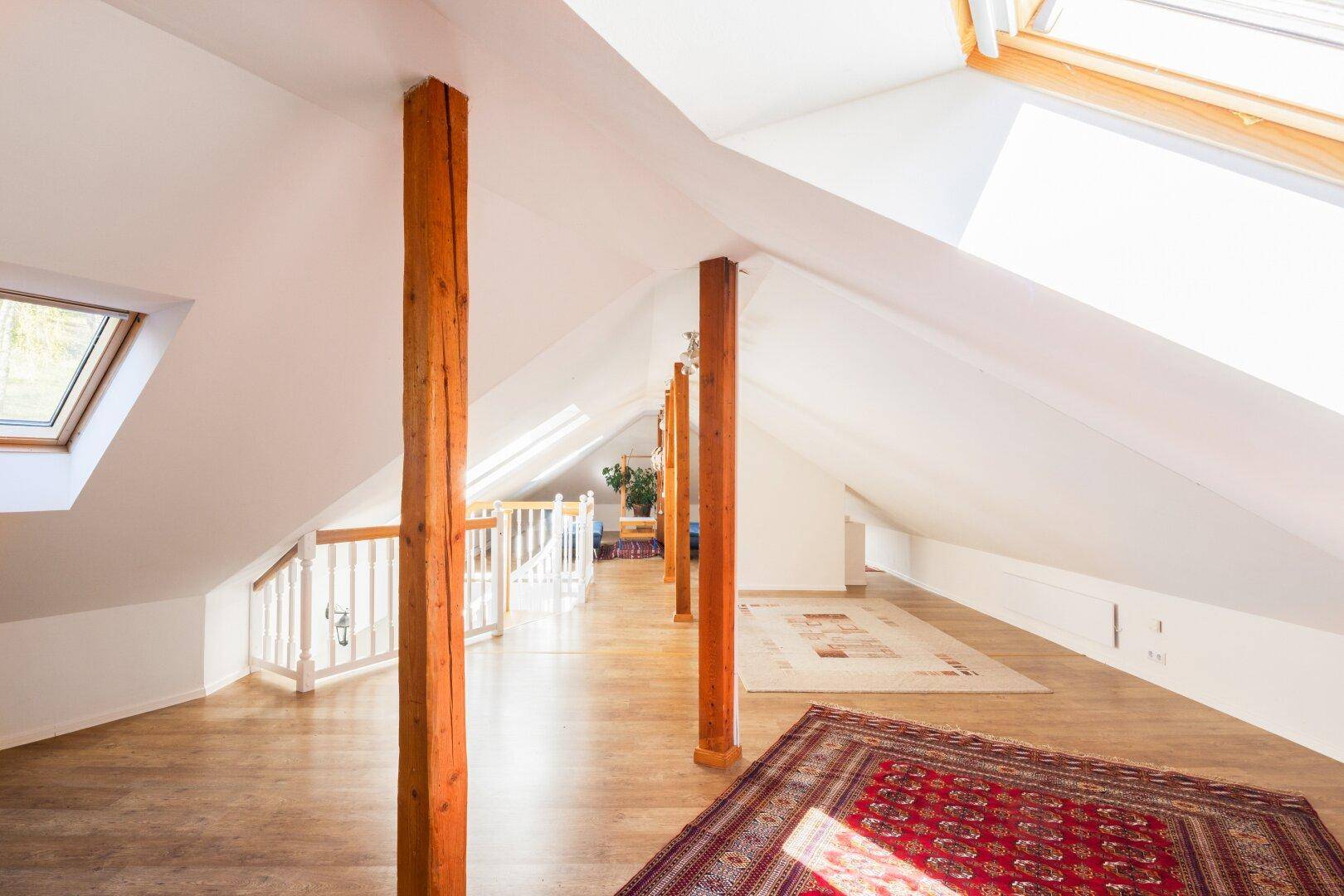 Geräumiges Dachgeschoss mit Holzbalken, Dachfenstern und Teppichen auf dem Holzboden.