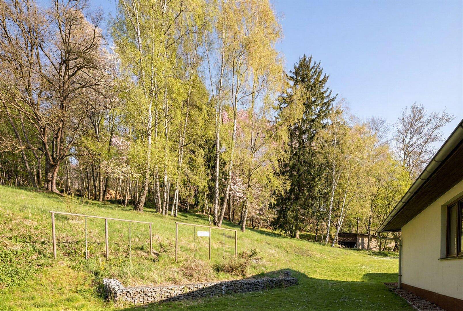 Seitenansicht des Hauses mit einer Gabionenwand und einem Hanggrundstück im Frühling.
