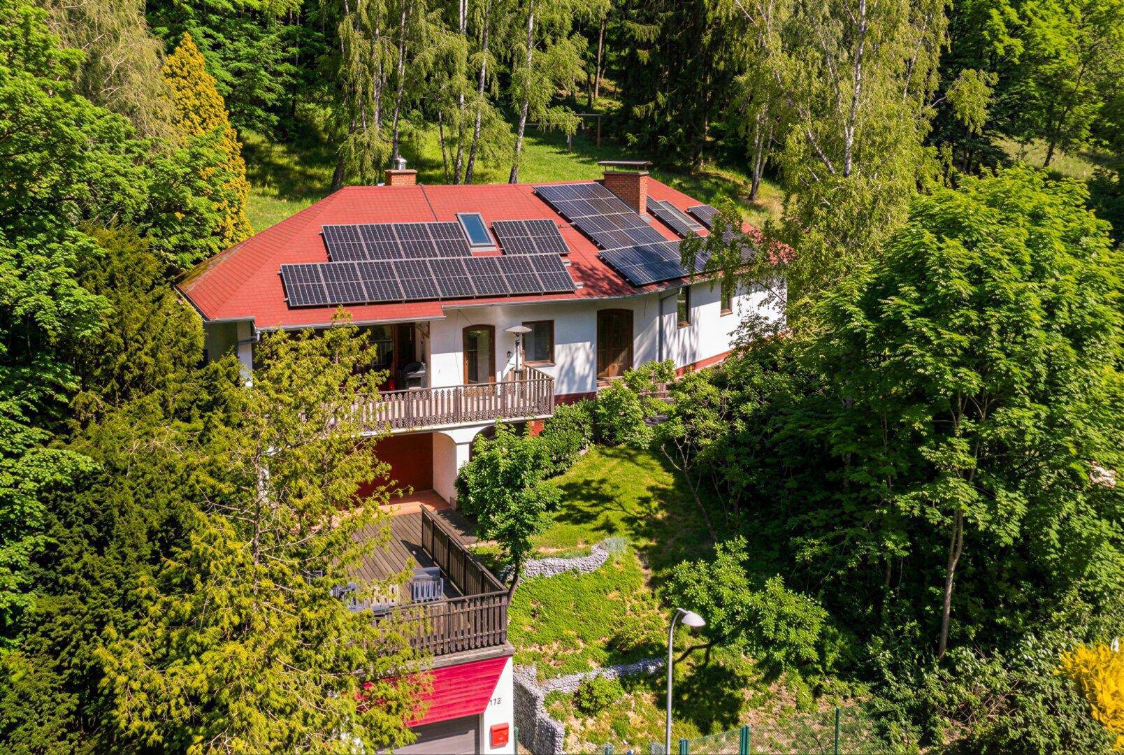 Haus mit rotem Dach und Solaranlage, eingebettet in eine grüne Hügellandschaft mit Bäumen.