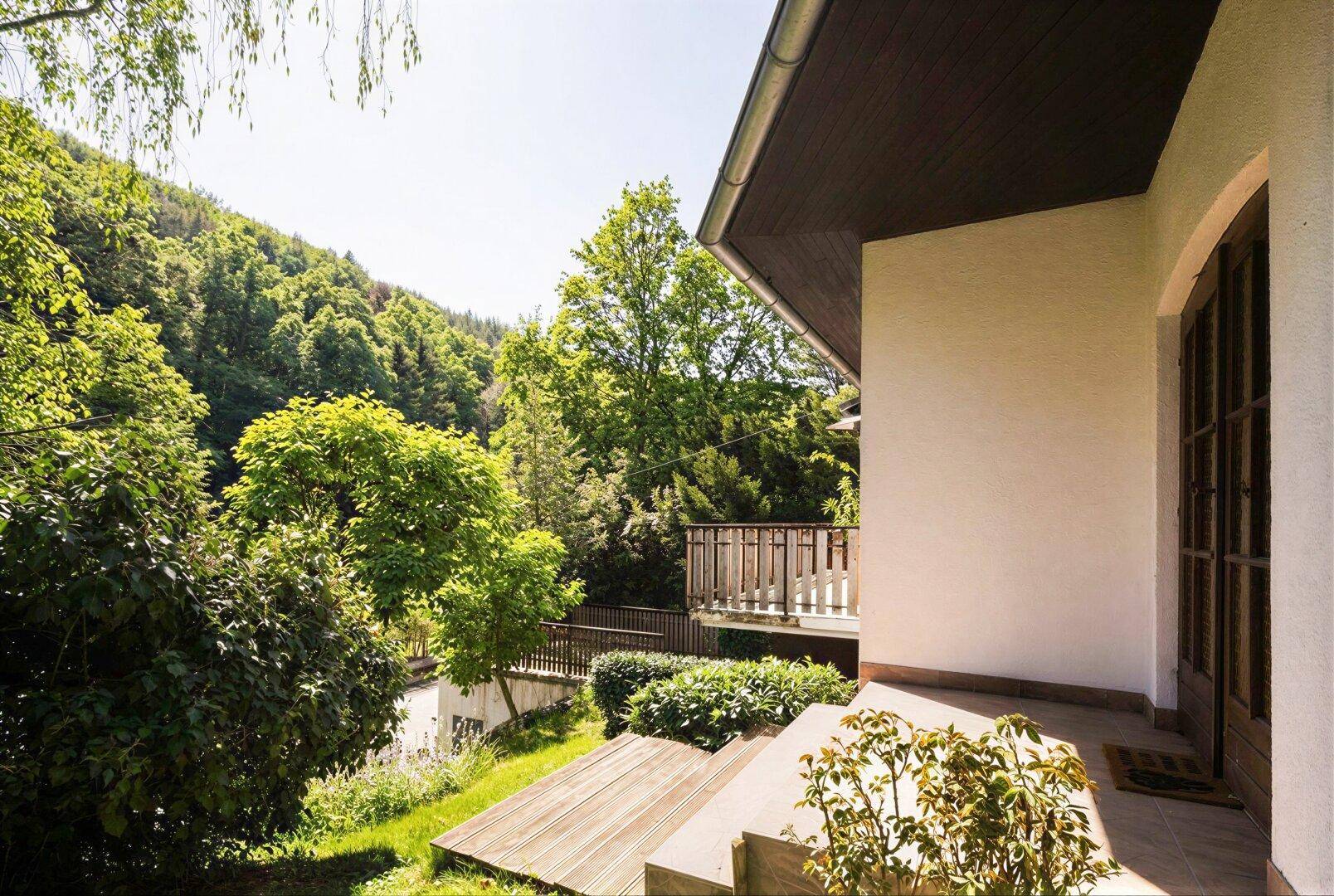 Seitlicher Blick auf das Haus mit Treppe und Holzboden, umgeben von viel Grün und Bäumen.