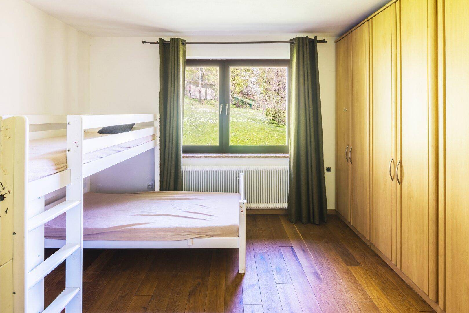 Helles Kinderzimmer mit Etagenbett, Holzschrank und Fenster mit Gartenblick.