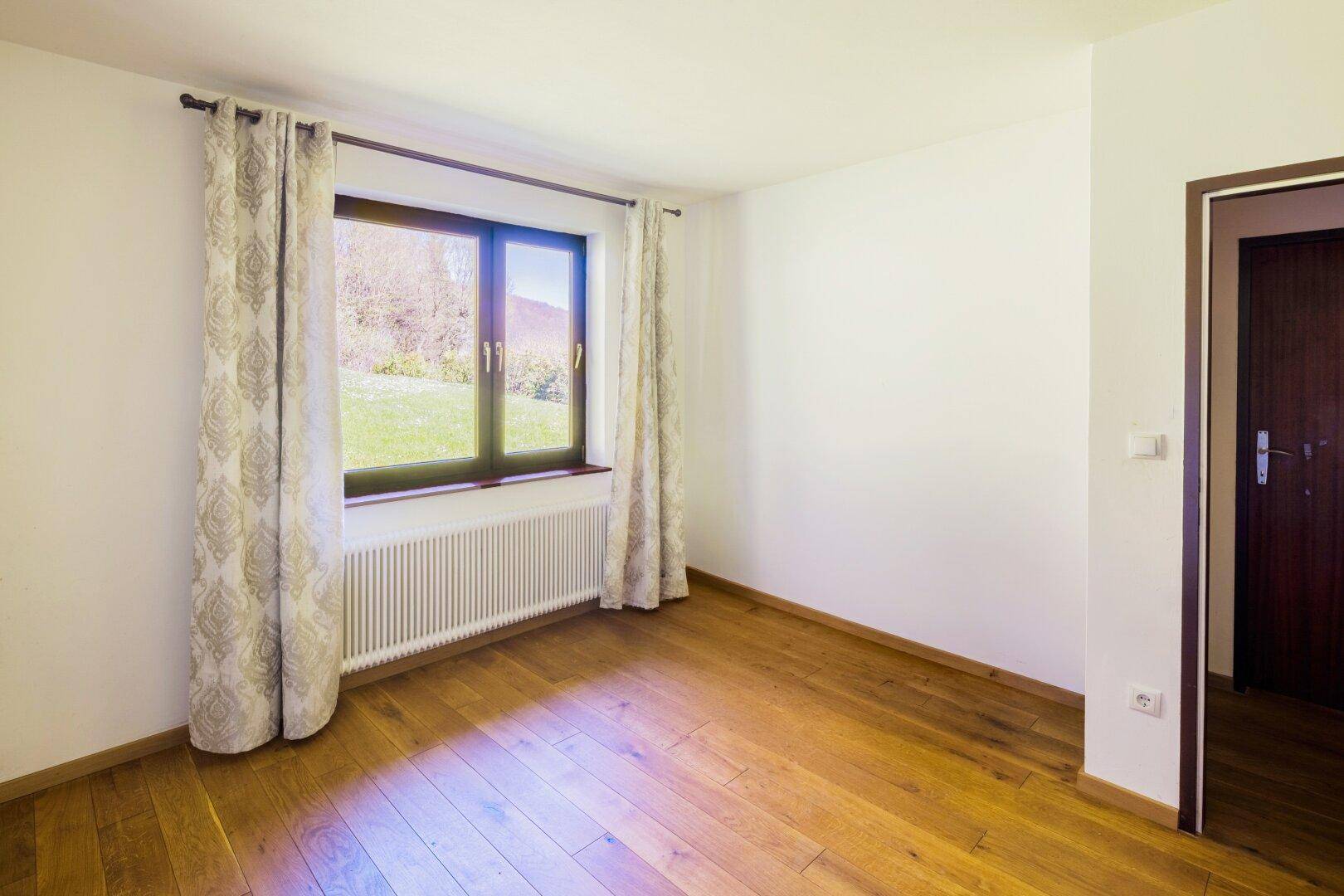 Leeres Zimmer mit hellem Holzparkettboden und Fenster mit Blick ins Grüne.