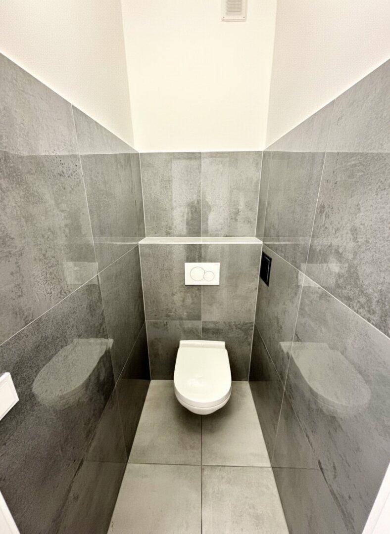 Separates WC mit modernen grauen Fliesen und wandhängender Toilette.