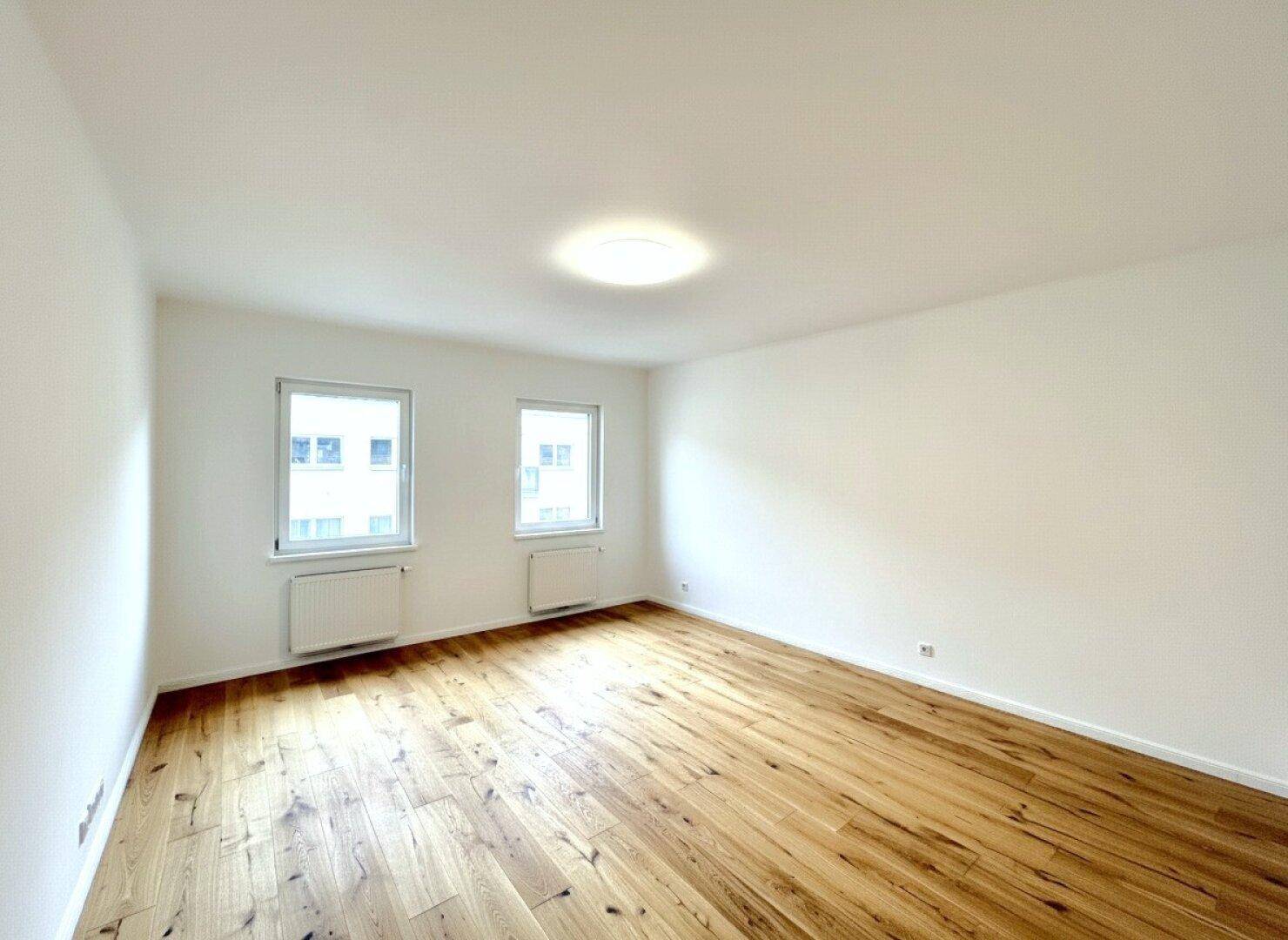 Geräumiger, heller Raum mit Parkettboden und zwei Fenstern, ideal als Wohnzimmer.