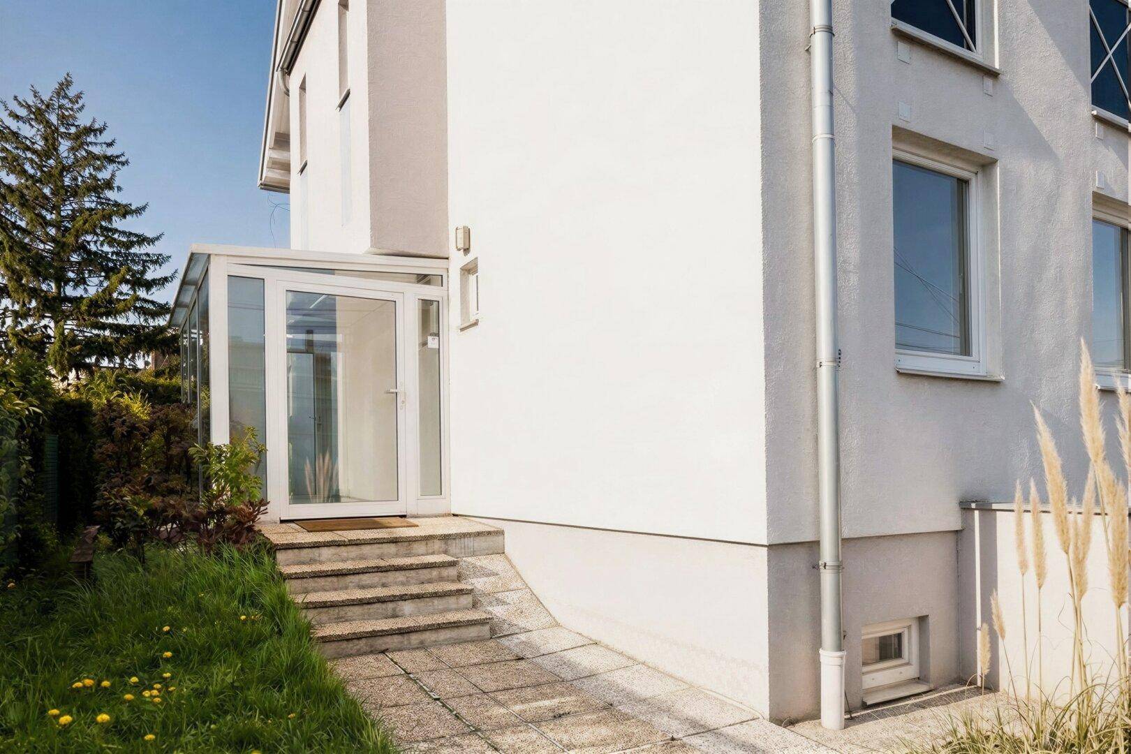 Moderne Hausfassade mit hellem Anstrich, verglastem Eingang und Zugang zum Untergeschoss.