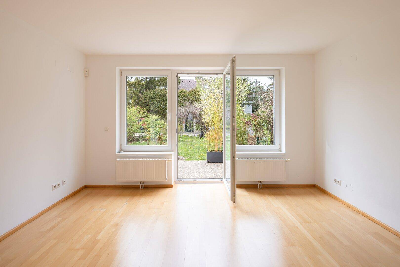 Heller Raum mit Holzfußboden und großen Fenstern, die Zugang zum Garten bieten.