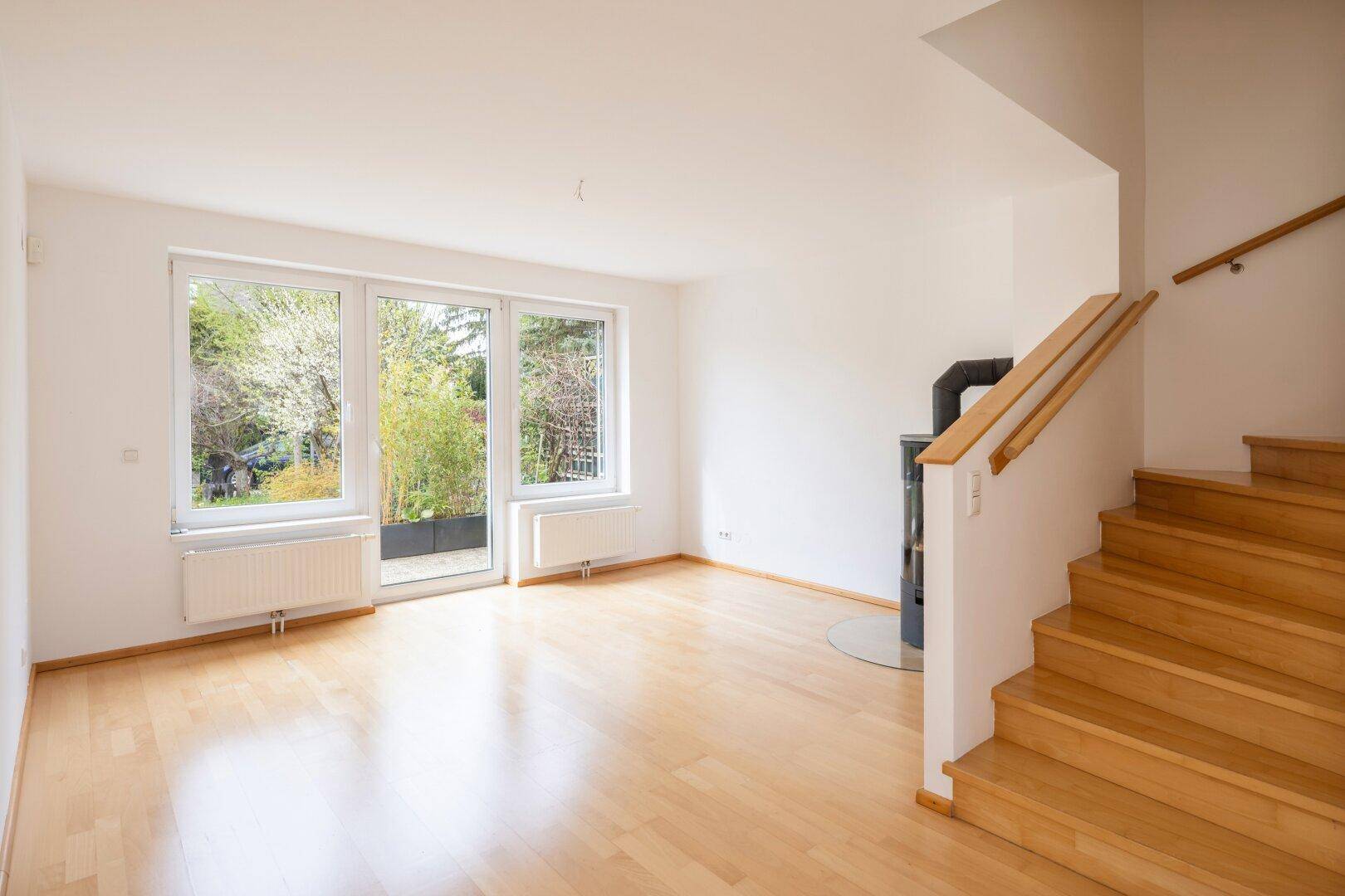 Heller Wohnbereich mit Kaminofen, Holzfußboden und großen Fenstern zum Garten.