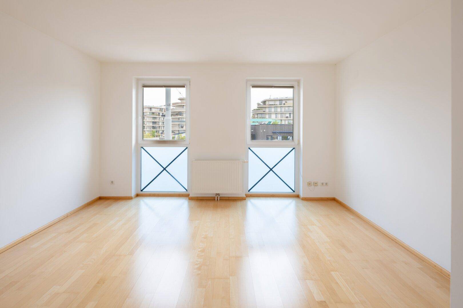 Helles, leeres Zimmer mit Holzfußboden und zwei Fenstern, ideal als Schlafzimmer.