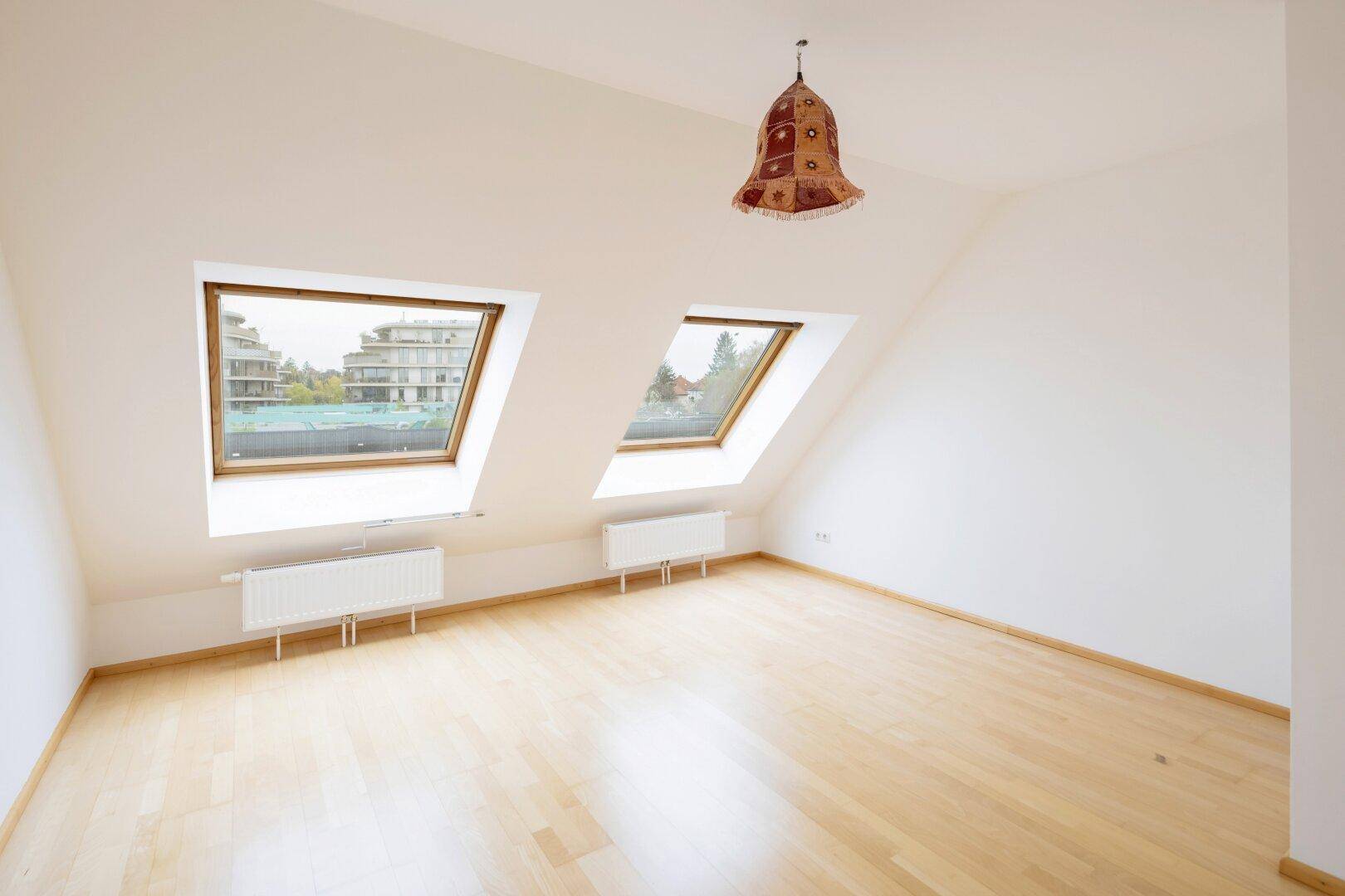 Dachzimmer mit schrägen Wänden, Holzfußboden und zwei Dachfenstern für viel Licht.