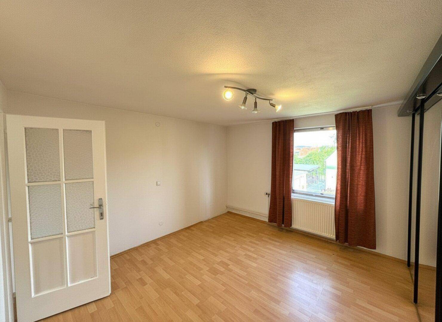 Helles Schlafzimmer mit Laminatboden und Fenster mit braunen Vorhängen.