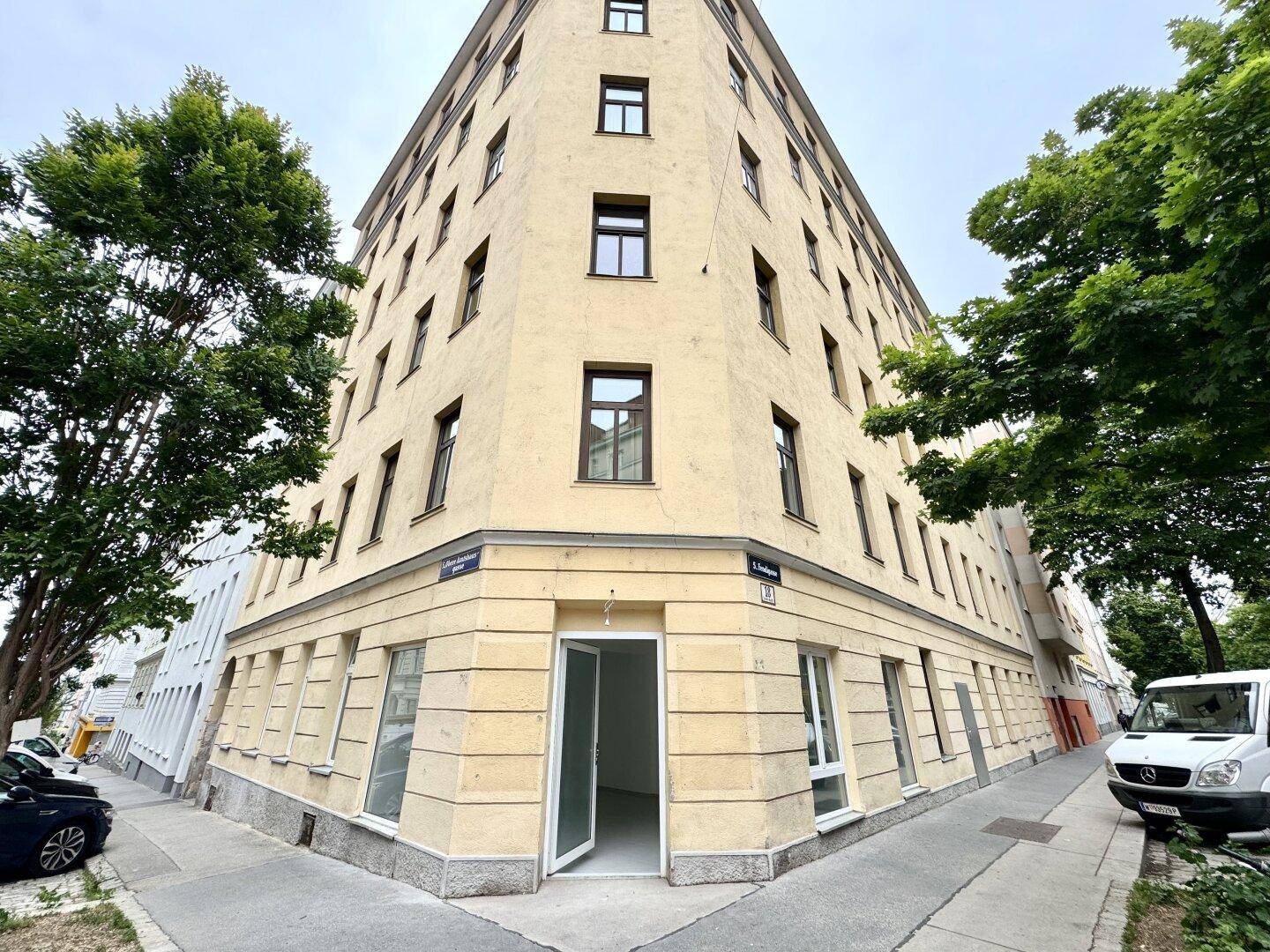 Eckgebäude mit gelber Fassade, vielen Fenstern und Bäumen, das eine städtische Umgebung zeigt.
