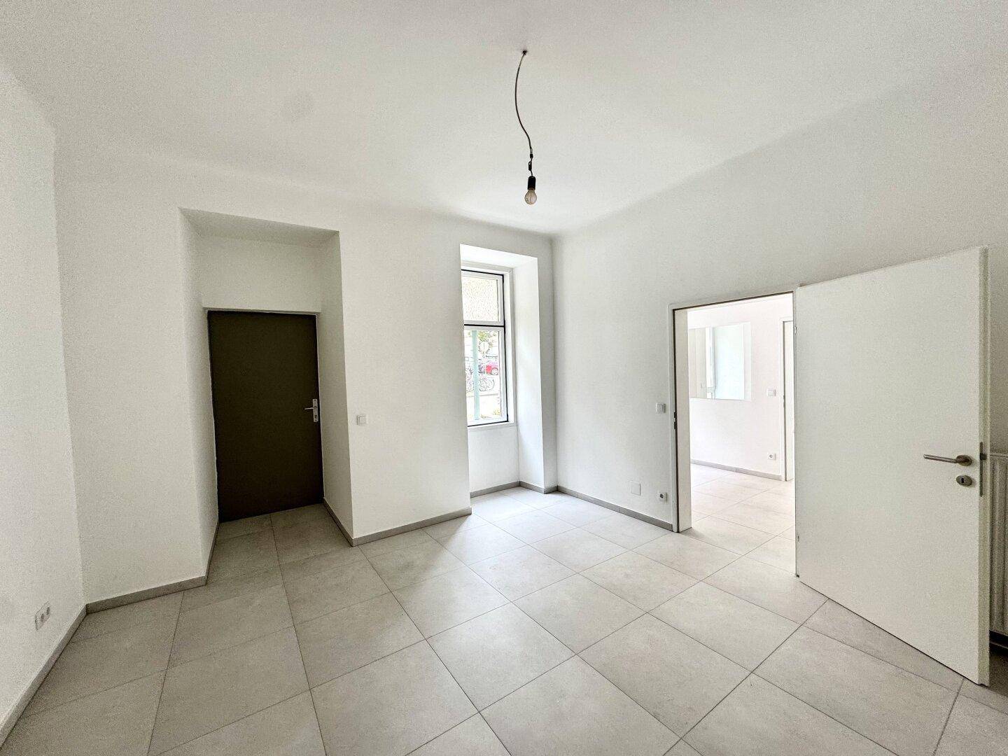 Heller Raum mit Fliesenboden, einem Fenster und einer dunklen Tür, bereit zur individuellen Nutzung.