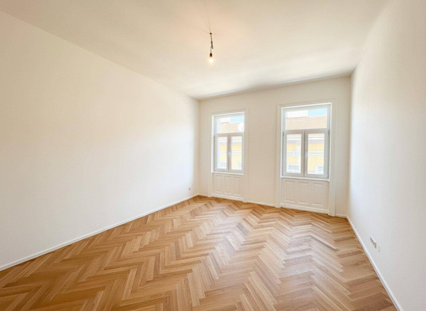 Heller Raum mit Fischgrätparkett und zwei Fenstern, ideal für ein Schlaf- oder Arbeitszimmer.