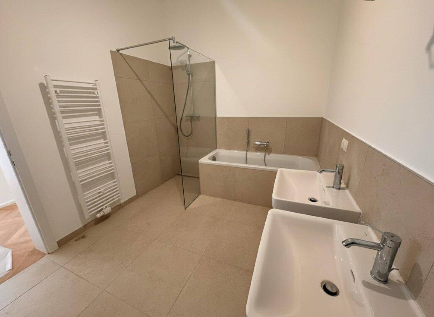 Helles Badezimmer mit Badewanne, Duschbereich, zwei Waschbecken und Fliesenboden.