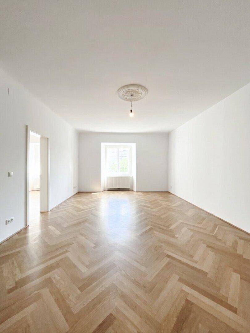 Geräumiges, leeres Wohnzimmer mit Fischgrätparkett und Stuckdecke, bereit für individuelle Gestaltung und Möblierung.