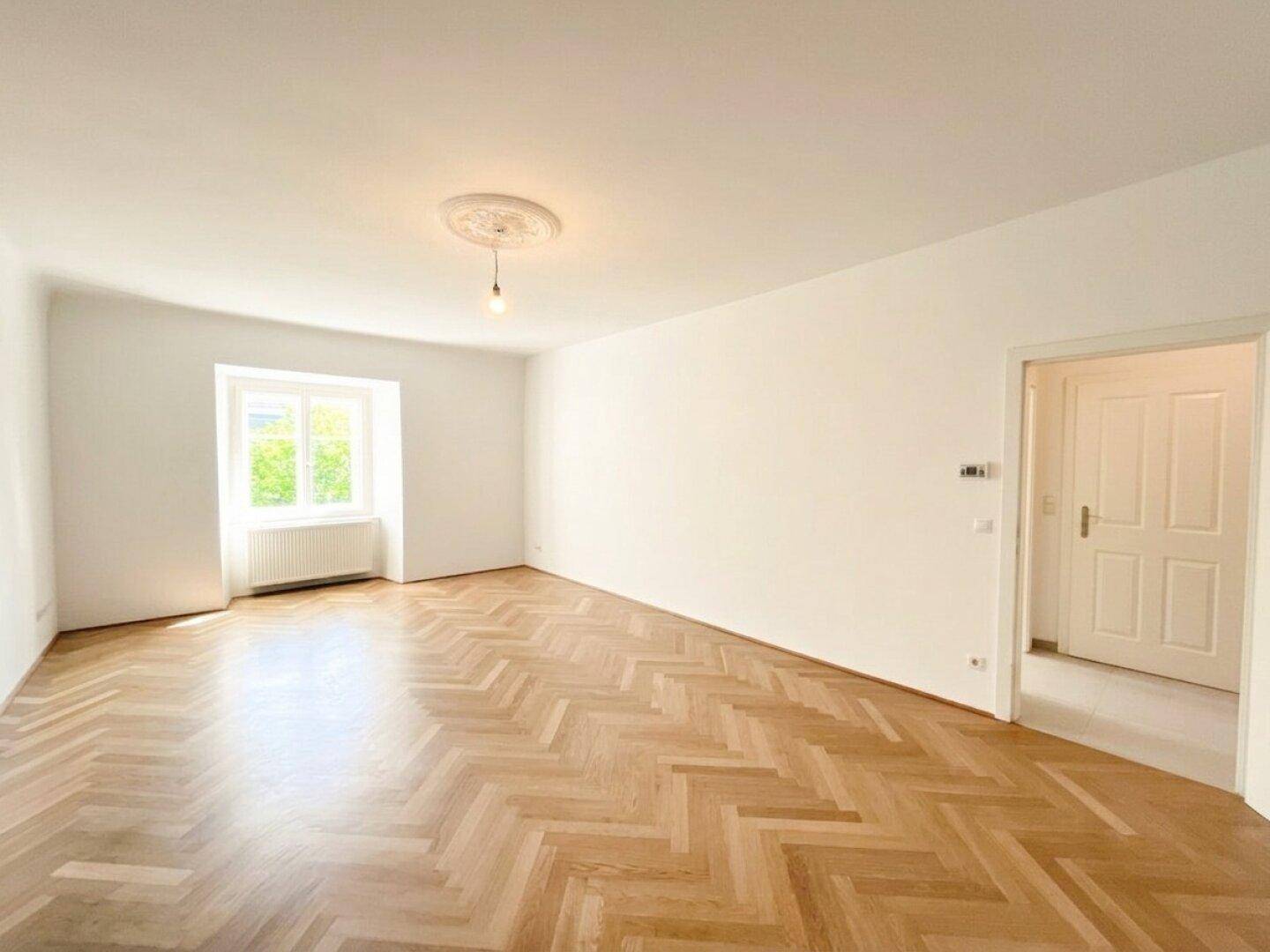 Lichtdurchflutetes Wohnzimmer mit Fischgrätparkett und großem Fenster, das viel Helligkeit in den Raum bringt.